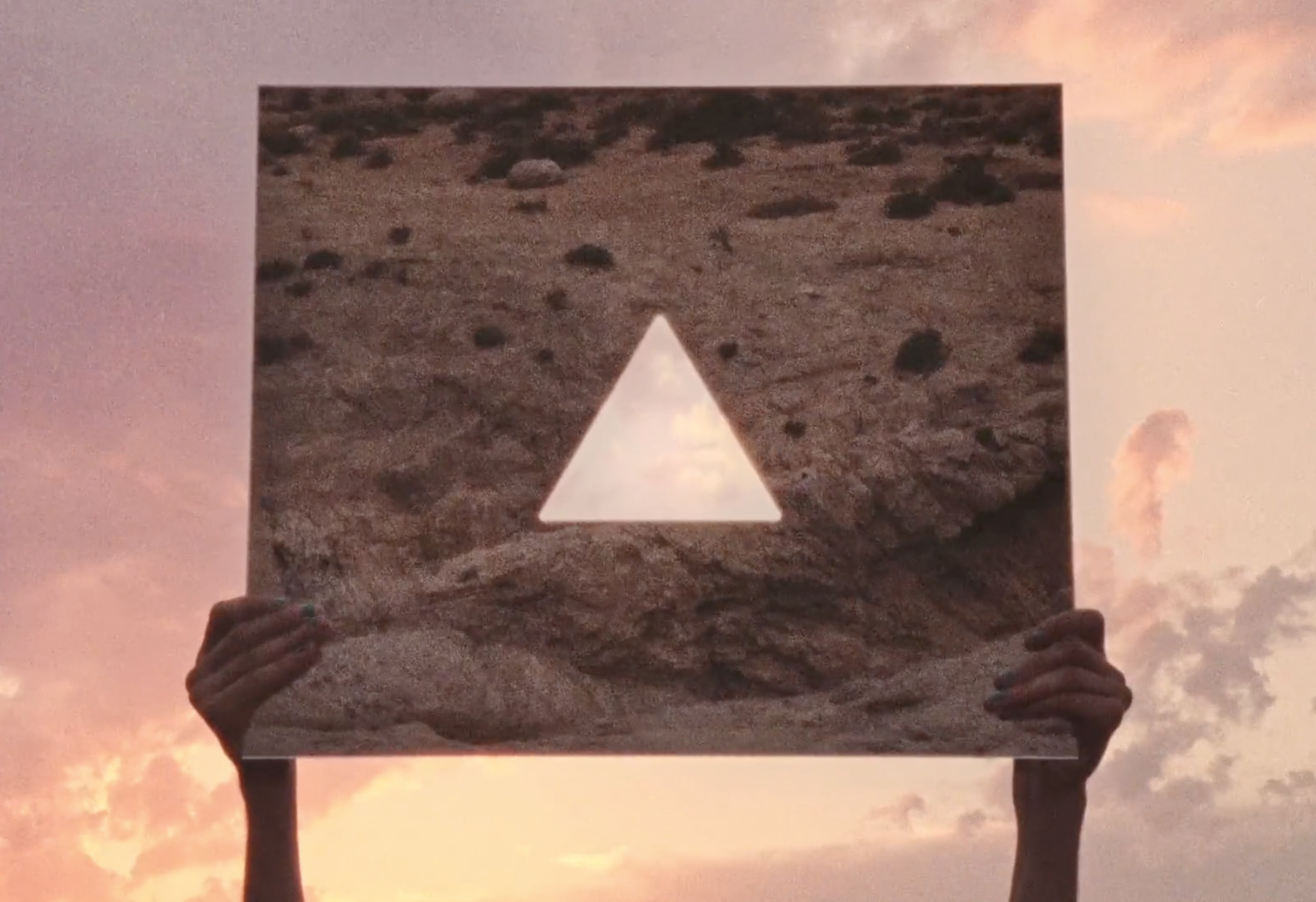 Someone holding up a photo of a desert landscape with a cut-out triangle in the center, against a sunset sky with clouds.