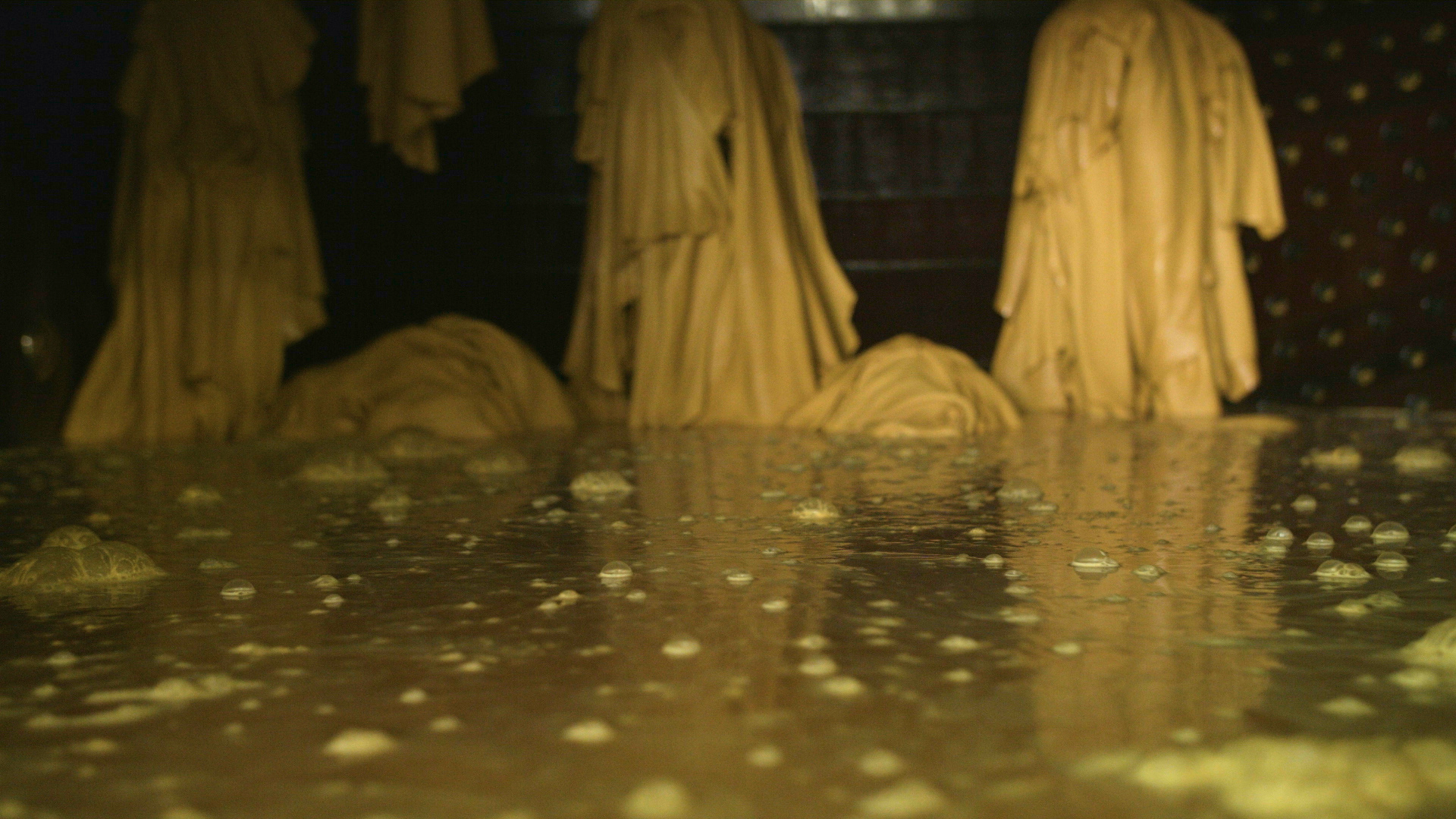 A close-up view of a gold-colored, textured surface with a reflective, shiny quality. The background features what appears to be draped fabric or paper with a tree bark or wood grain pattern, and some dark areas with small, circular patterns.
