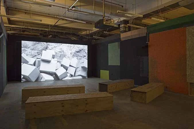 An empty room with wooden benches, a large screen displaying a black-and-white image of tilted cubes, and colorful geometric wall panels, possibly a gallery or presentation space.