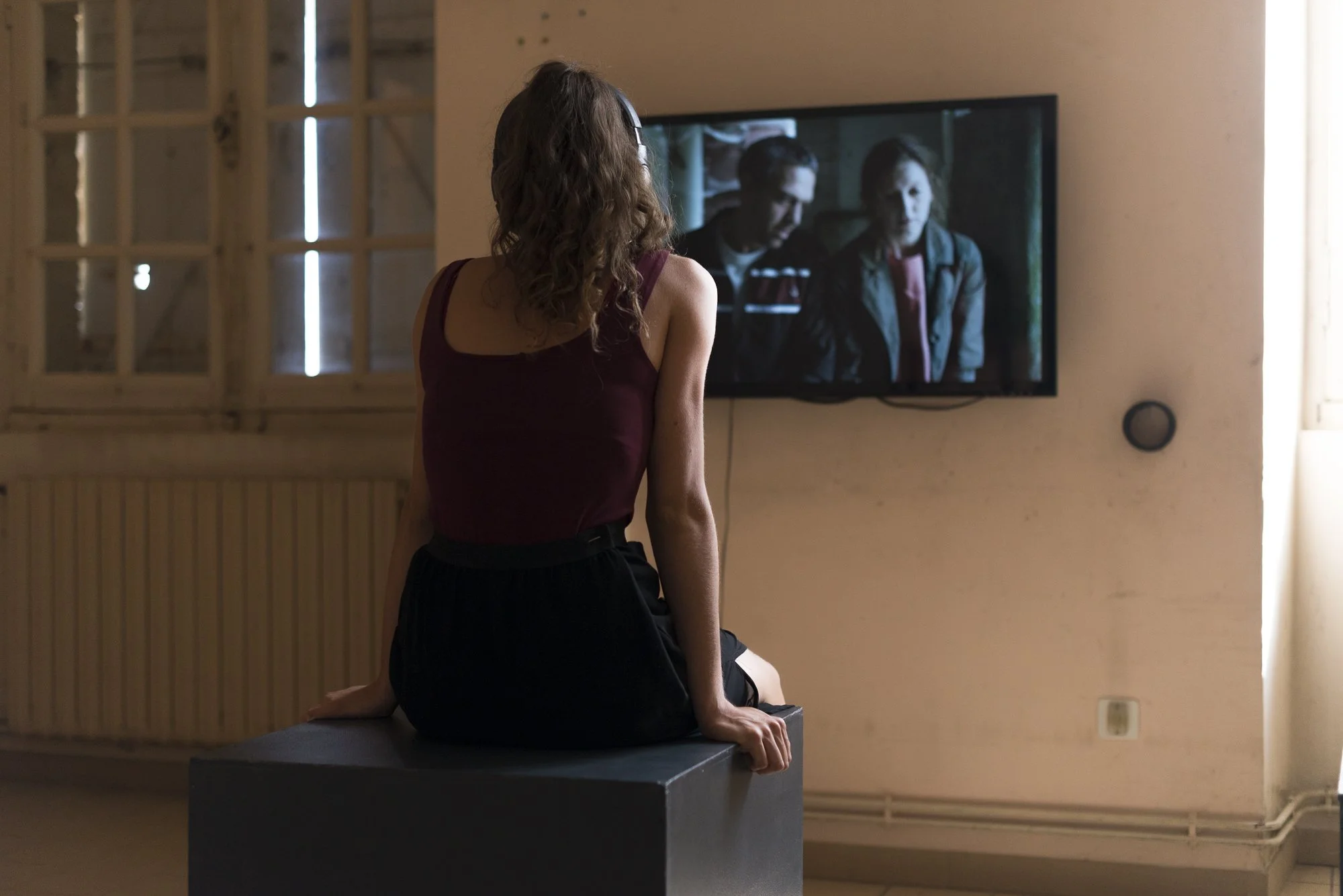 A woman with curly hair sitting on a black bench watching a TV screen in a room with beige walls and a window.
