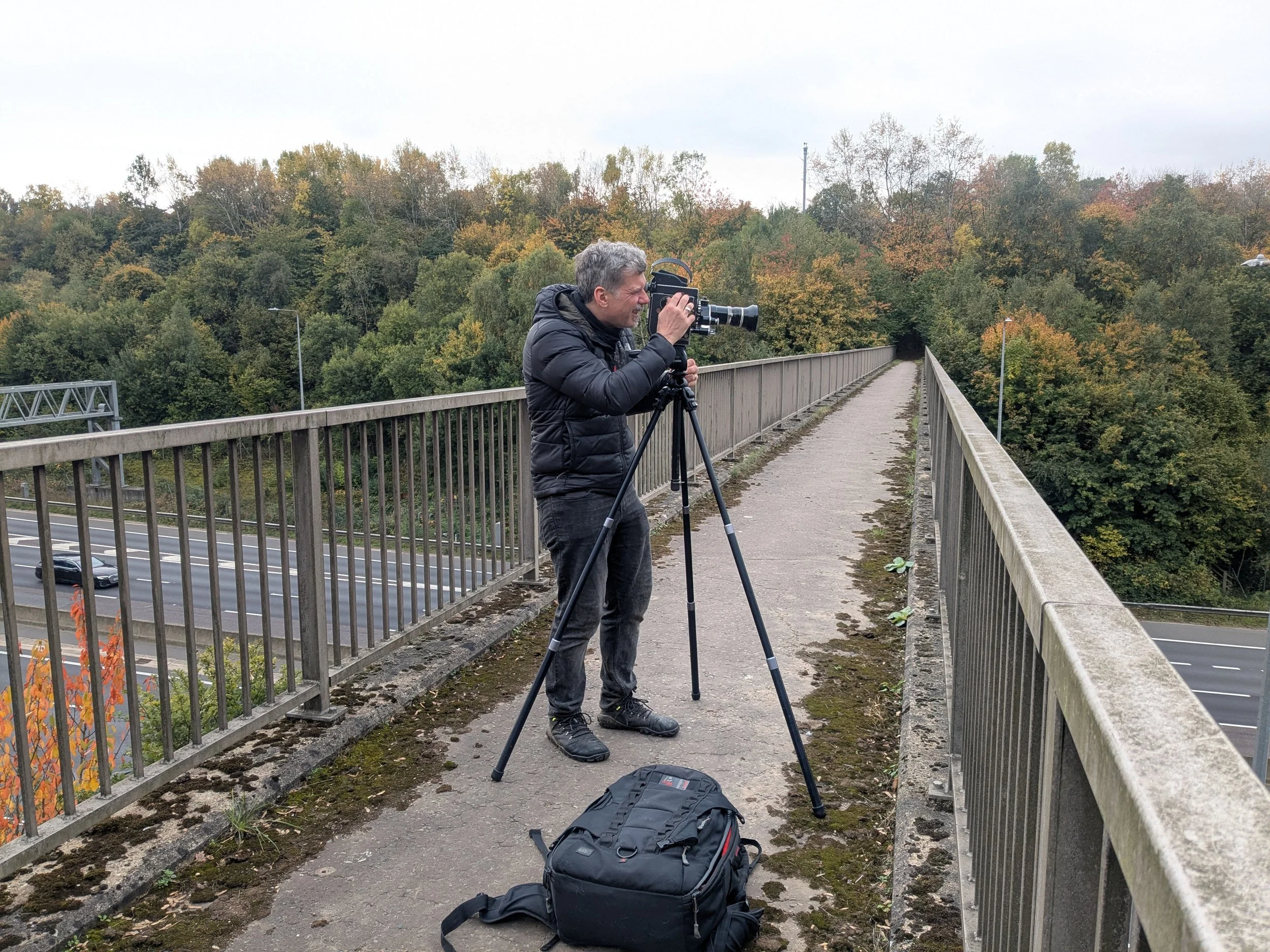 A man in a black jacket and pants operating a camera on a tripod on a pedestrian bridge over a highway surrounded by trees with autumn foliage.