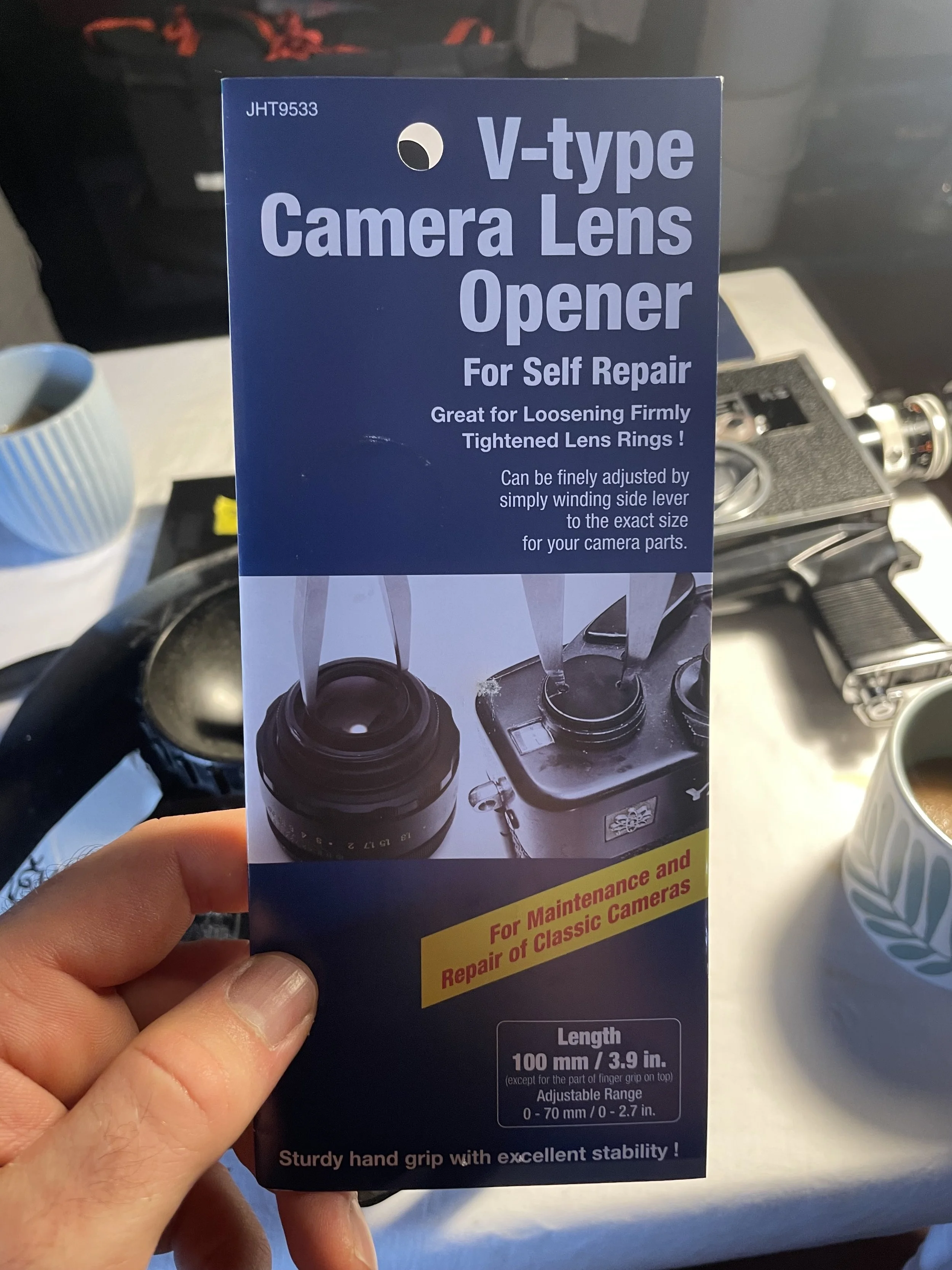 Packaging for a V-type camera lens opener, designed for self-repair of vintage camera lenses. The package indicates it is used for loosening and tightening lens rings and can be adjusted by winding a side lever. It features a sturdy hand grip and is 