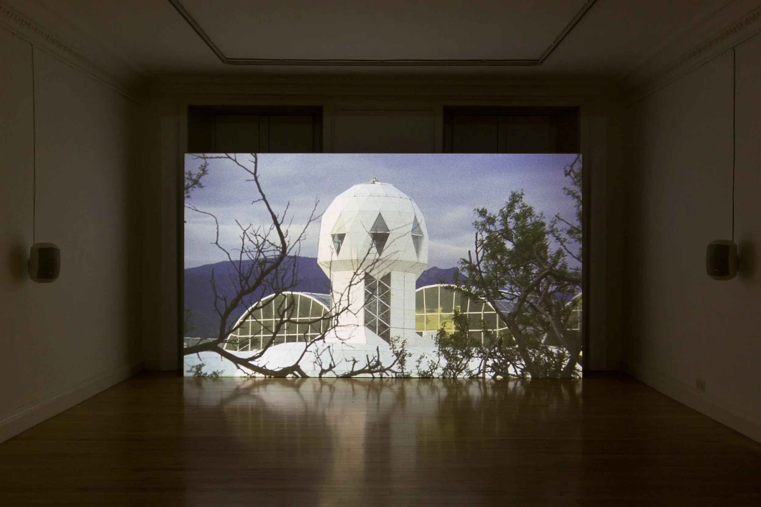 Indoor gallery with a projected image of a building featuring a geodesic dome, trees, and mountains in the background.