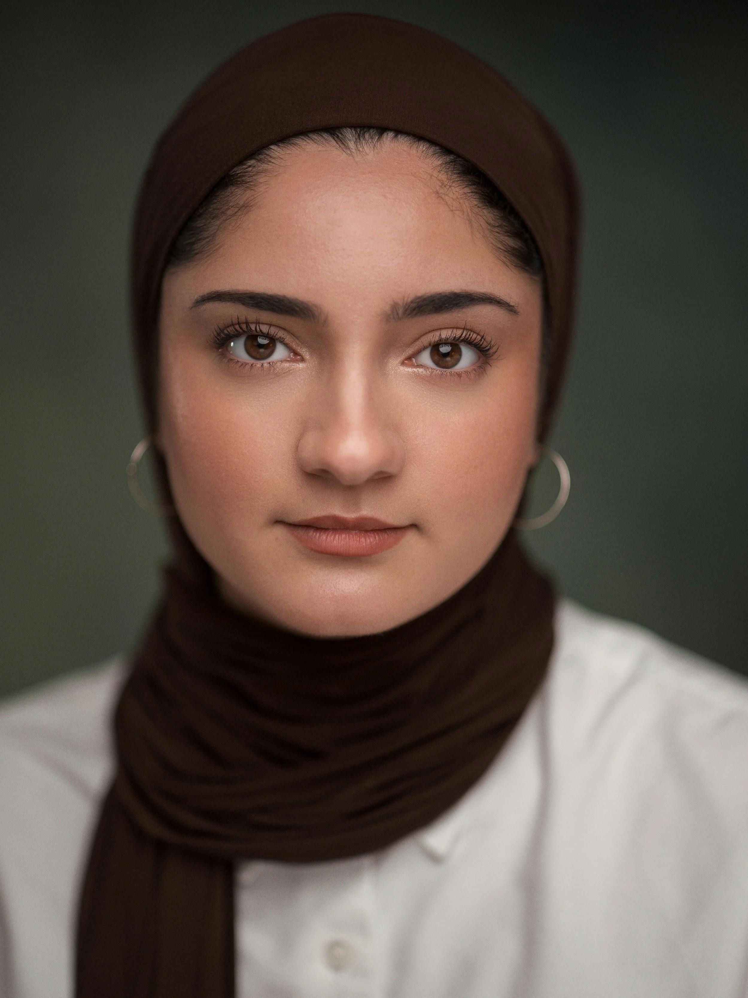 Close-up portrait of a young woman with light brown eyes, wearing a dark brown headscarf and hoop earrings, against a blurred dark green background.