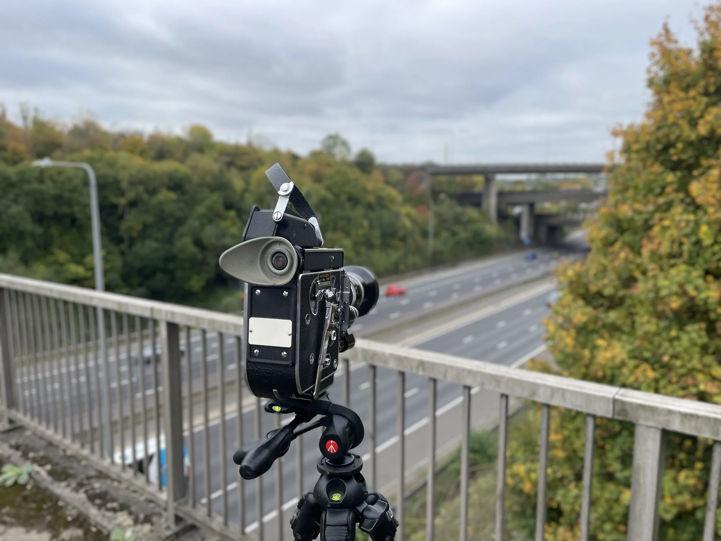 Tripod-mounted surveillance camera overlooking a highway with cars and trees, under a cloudy sky.