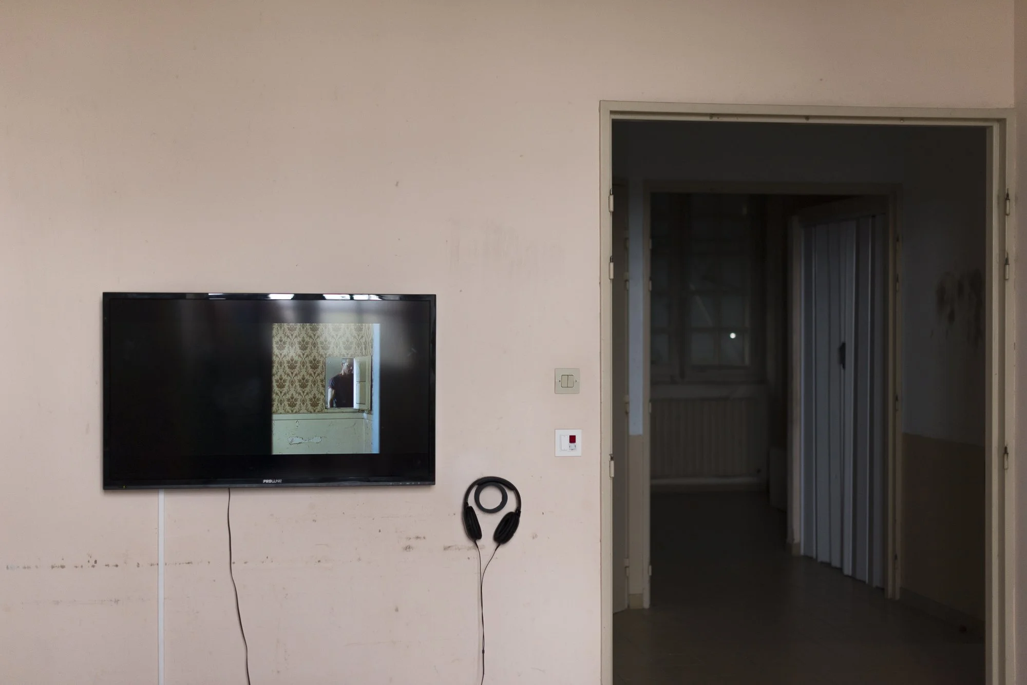 A living room with a wall-mounted TV displaying a mirror reflecting a person, a pair of black headphones hanging on the wall, and an open bathroom door leading to a dimly lit room.