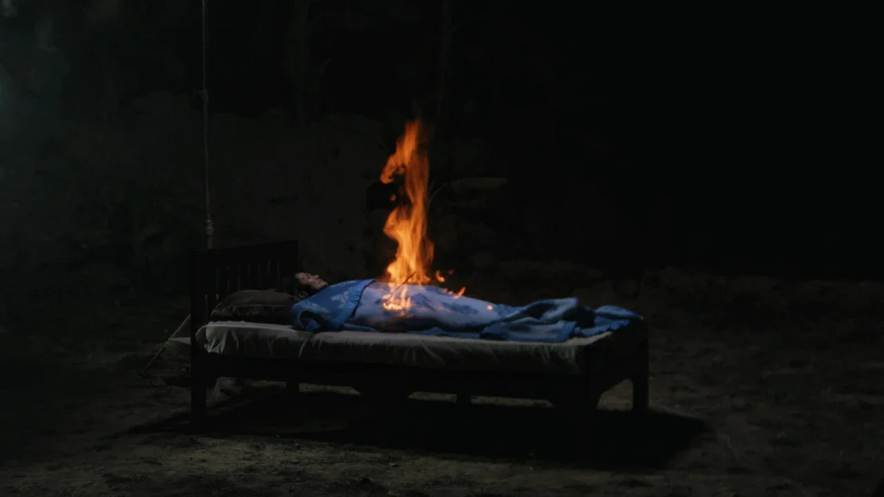 Person lying on a bed outdoors at night with flames rising from their midsection.