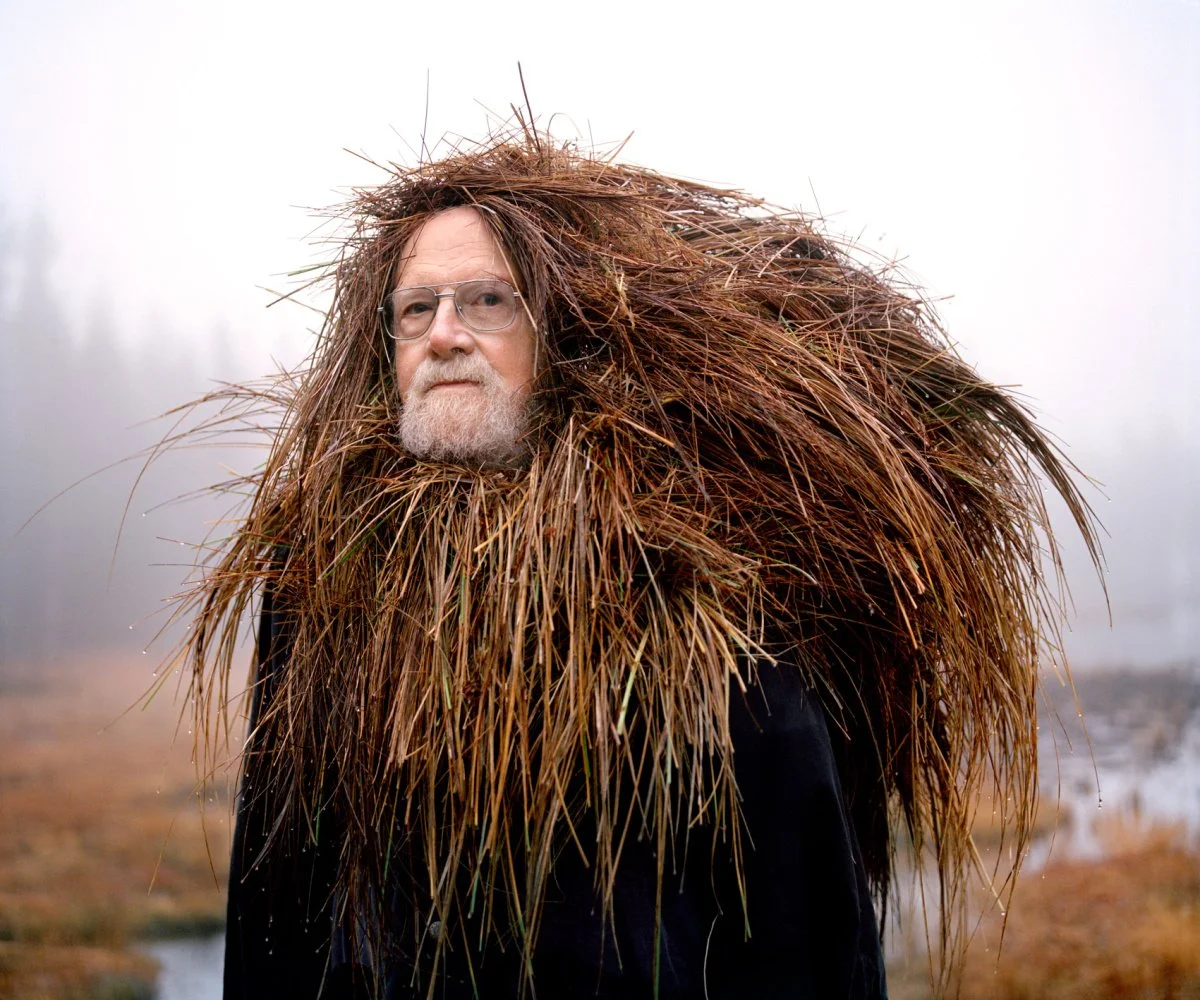 Man with glasses and a beard wearing a large, bushy coat made of brown dried grass or leaves, standing outdoors in a foggy, marshy landscape.