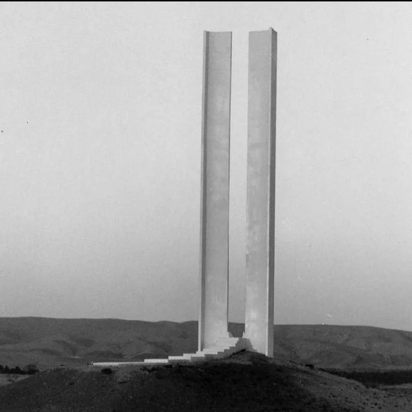 Two tall, vertical concrete monuments with curved steps at the base, set against a plain, cloudy sky and hilly landscape.