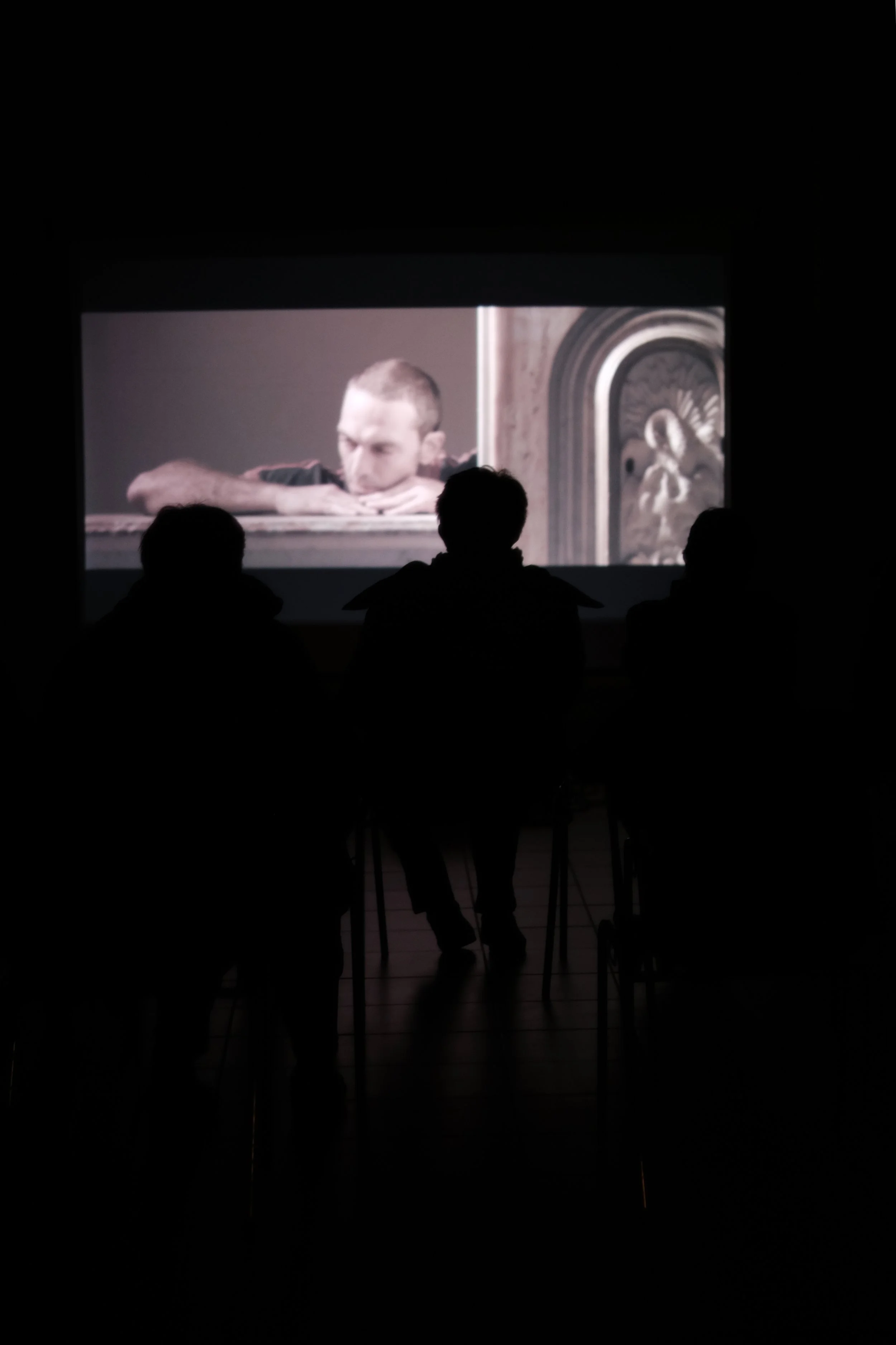 People watching a movie in a dark theater with the screen showing a man resting his head on a surface, with a religious or historical painting visible on the right side of the screen.