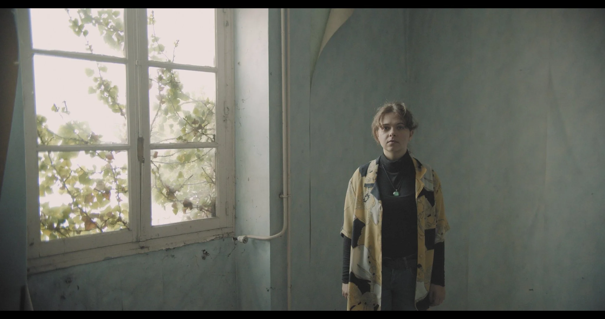 Young woman with short, wavy hair standing in an old, worn room with a large window and green foliage outside. She is dressed in a patterned yellow and black jacket, black turtleneck, and wears a necklace with a green pendant.