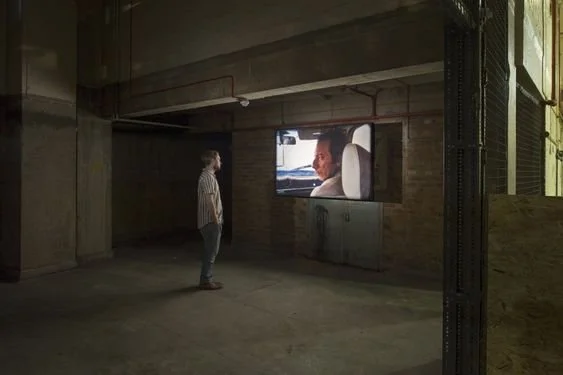 A person stands alone in a large, empty industrial-style room, watching a large screen displaying a woman on a video call or TV, with concrete walls and minimal lighting.