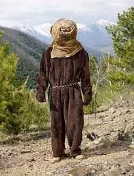 Person wearing a brown jumpsuit and wide-brimmed hat walking on a dirt trail in a mountainous forest area.