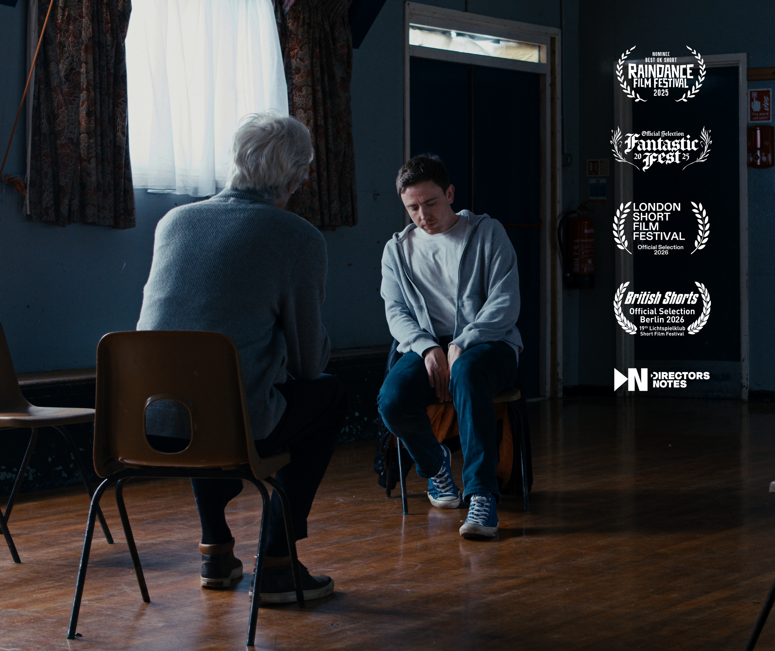 the-parish-raindance-film-production-st-albans.jpg A man sitting looking down with a sad expression, an older man, seen from behind, sits facing him in a room with wooden floors and worn walls. There are awards for film festivals on the right side. 