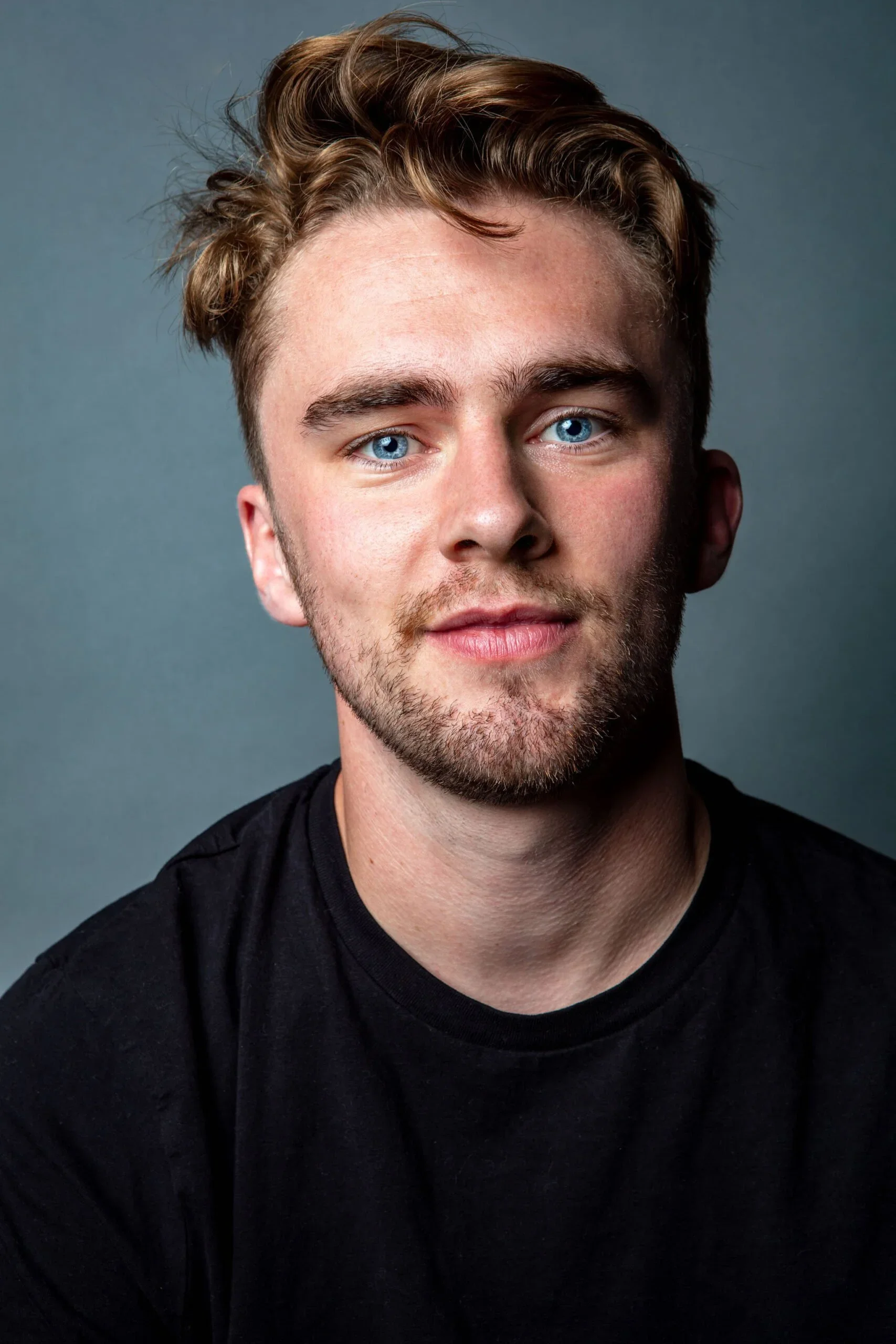 Close-up portrait of a young man with light skin, blue eyes, light brown hair, and a beard, wearing a black shirt, smiling slightly, set against a gray background.