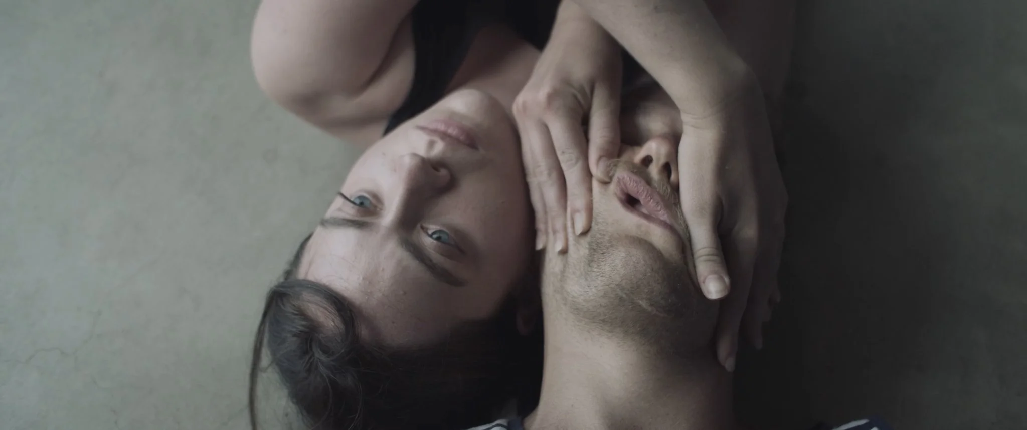 Two people lying on the floor, one with eyes open and one with eyes closed, holding each other's faces in an intimate pose.