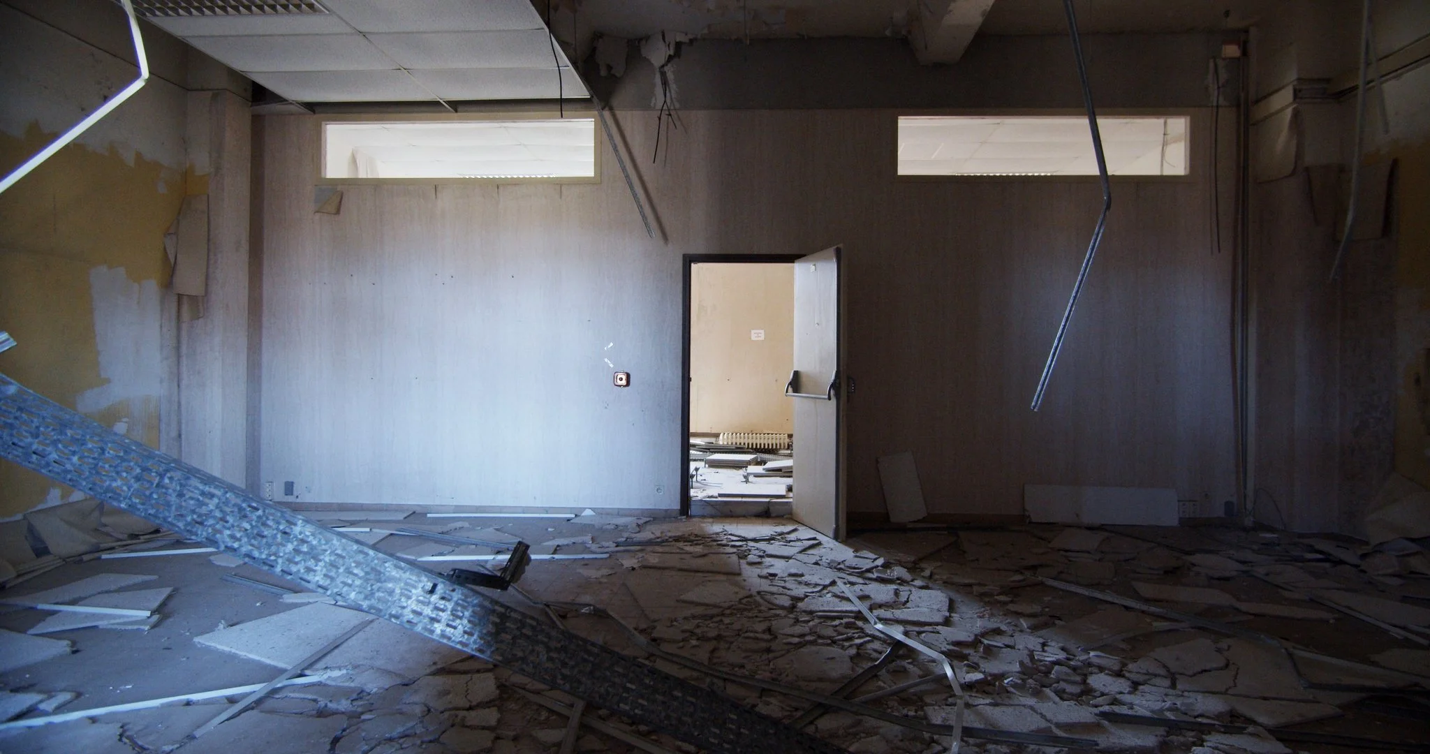 Interior of a damaged room with debris on the floor, exposed ceiling beams, broken drywall, and an open door leading to another room.