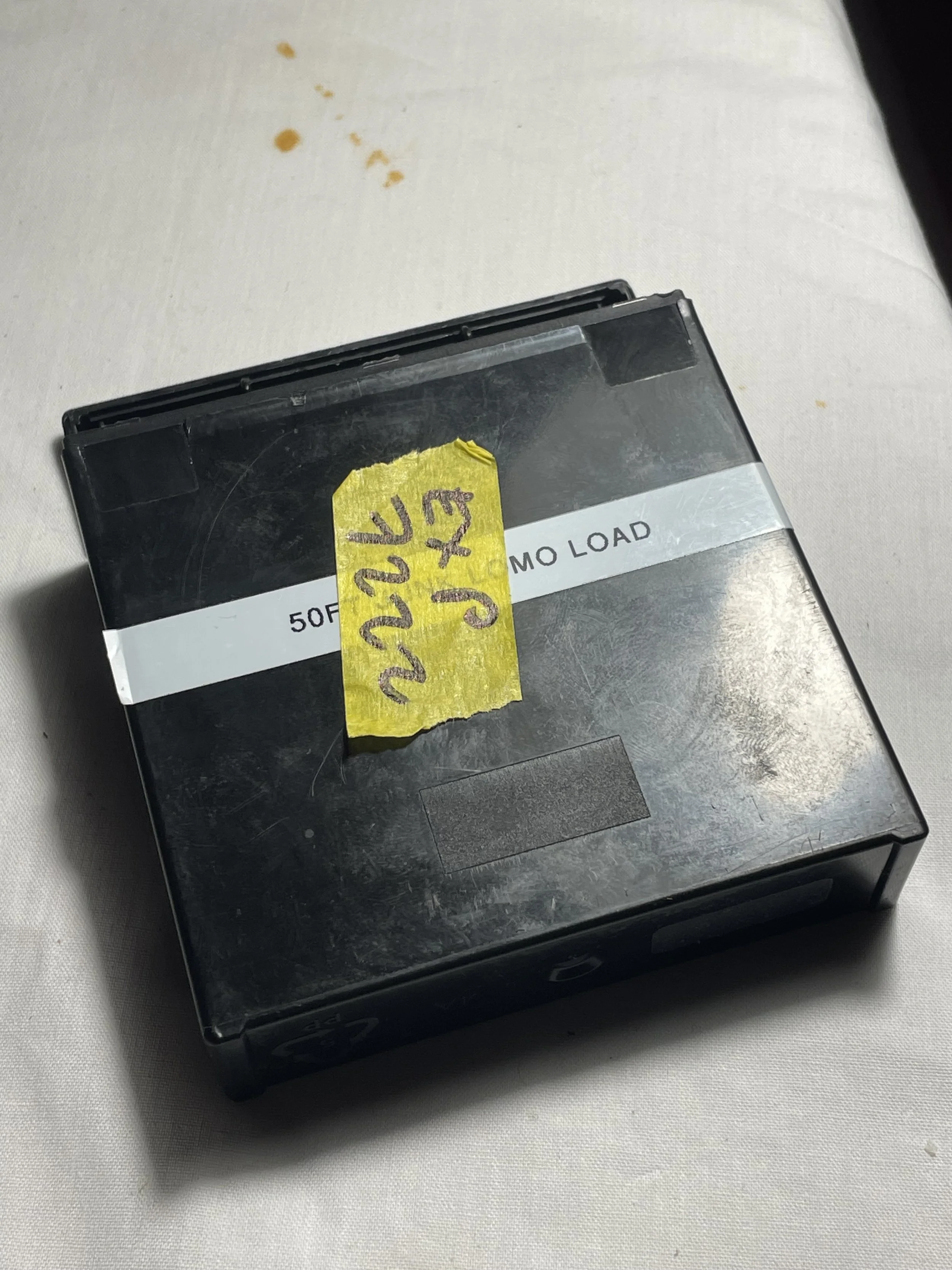 Black 50-pack film or photo paper box with a white label and yellow tape on top, placed on a white surface with small brown stains.