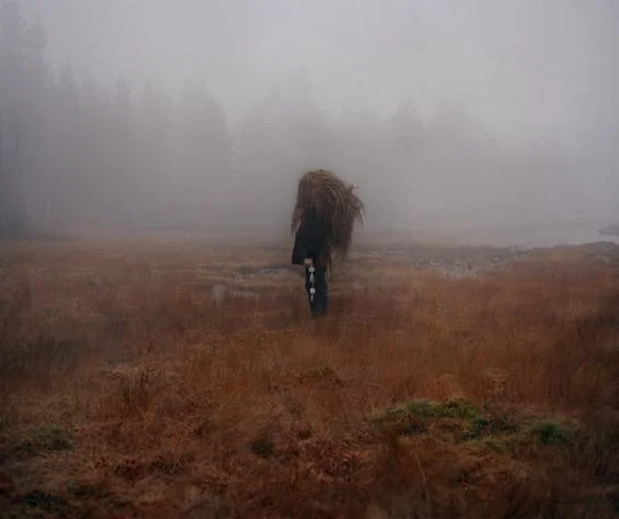 A person walking through a foggy, open field with tall grass, carrying a large bundle or load on their back.