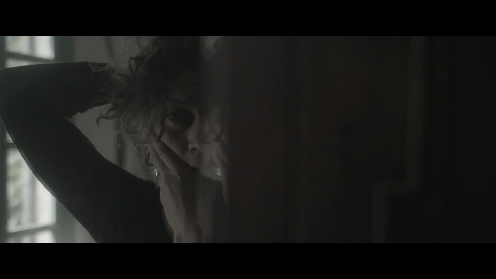 A woman with curly hair resting her face on her hand, looking at the camera through a reflective surface with a window in the background.