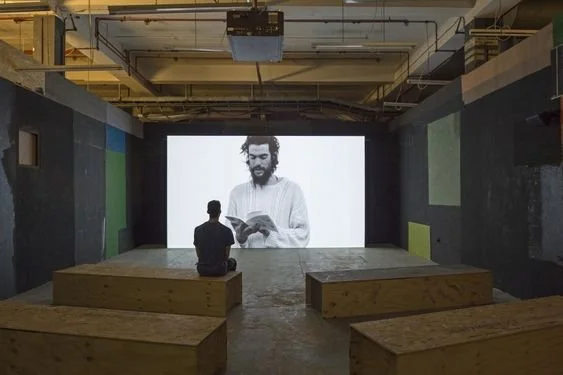 Person sitting on a wooden bench in front of a large digital screen displaying an image of a bearded man reading a book inside a dark industrial room.