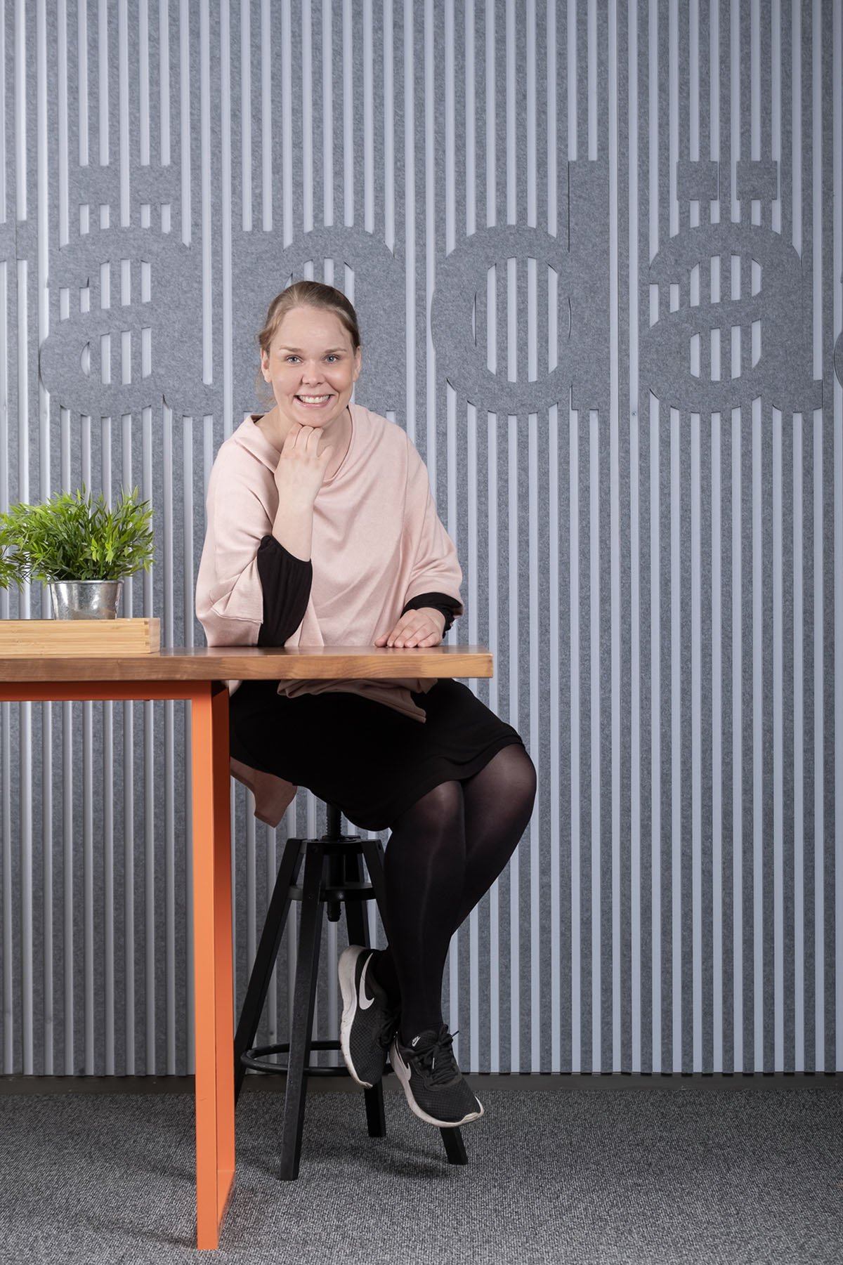 Nainen istuu jakkaralla, hymyilee, taustalla brändäämön logo seinässä.