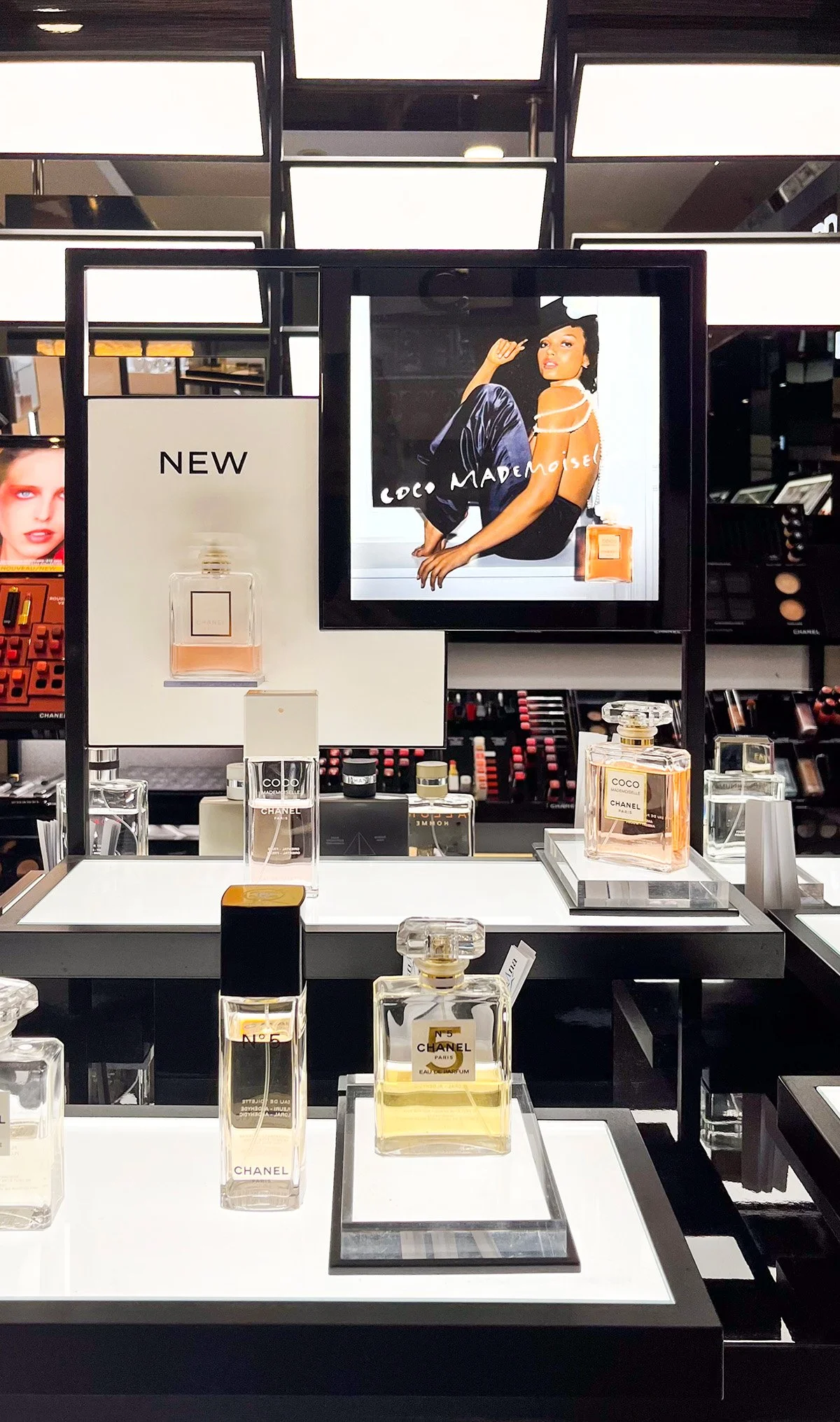 Lähikuvassa Chanelin shopista Chanel N°5 -tuoksu ja Coco Mademoiselle -tuoksu, taustalla digitaalinen mainosnäyttö ja muuta kosmetiikkaa.