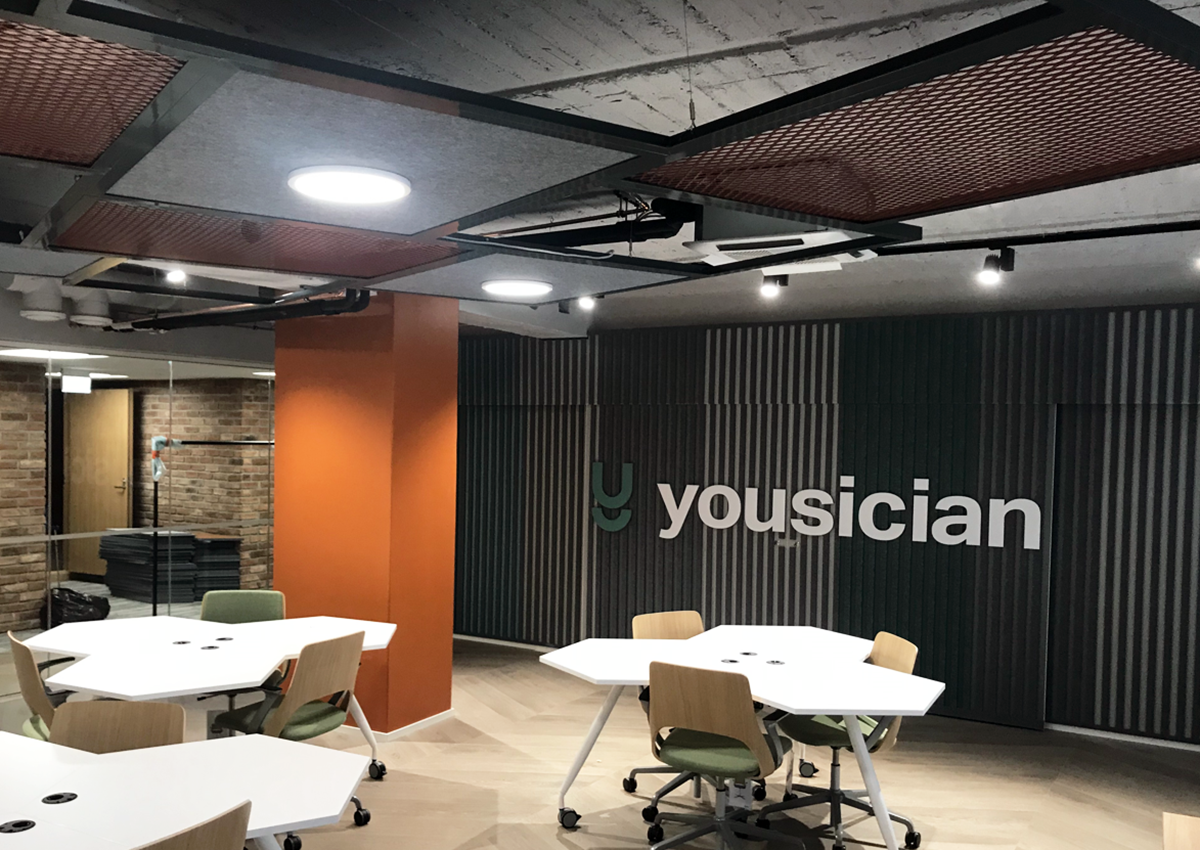 Tilassa näkyy työtila, jossa on pöytiä ja tuoleja, seinällä logo 'yousician', katto ja seinät on sisustettu modernisti.