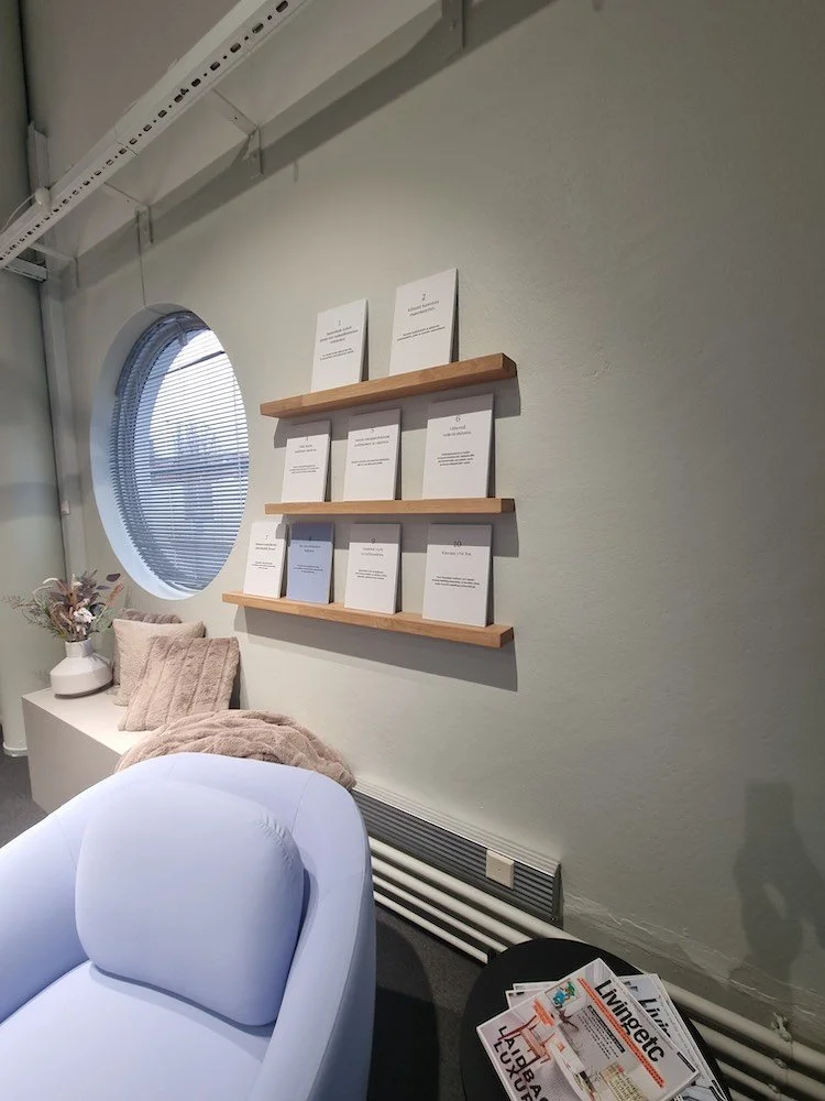 Näkymä sisätiloista, jossa on valkoinen nojatuoli, pöytä, lattiatyynyjä, seinällä puiset hyllyt ja kirja- ja lehtikasat, sekä pyöreä ikkuna.