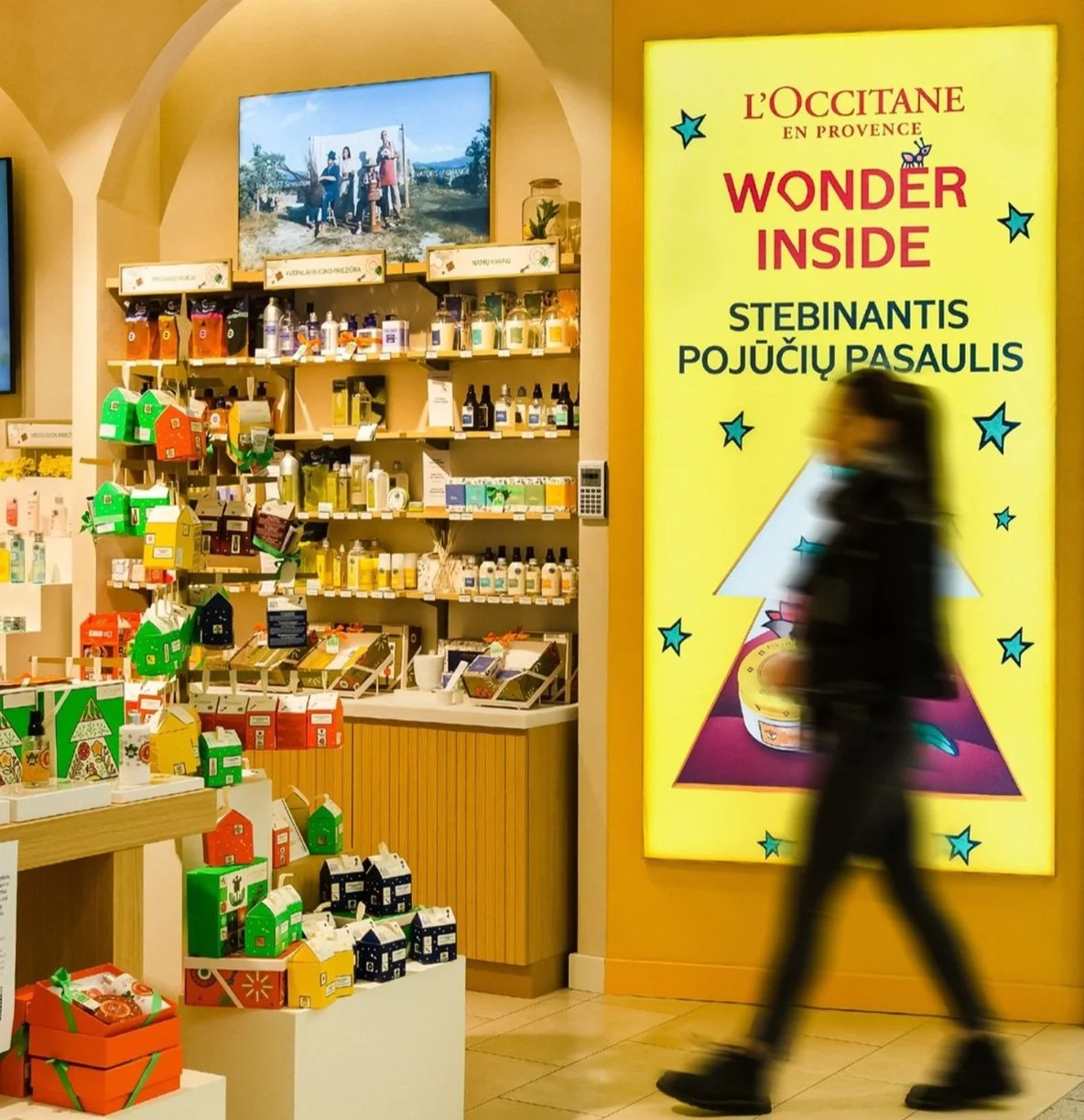 Myymälä, jossa on kosmetiikkatuotteita ja kuvassa keltainen led paneelinäyttö kävelevän naisen takana. 