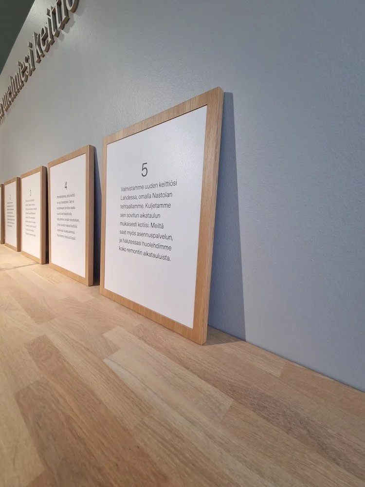 Taulut vaalealla puisella kehyksellä seinää vasten rivissä. Tauluissa on tekstiä suomeksi valkoisella pohjalla.