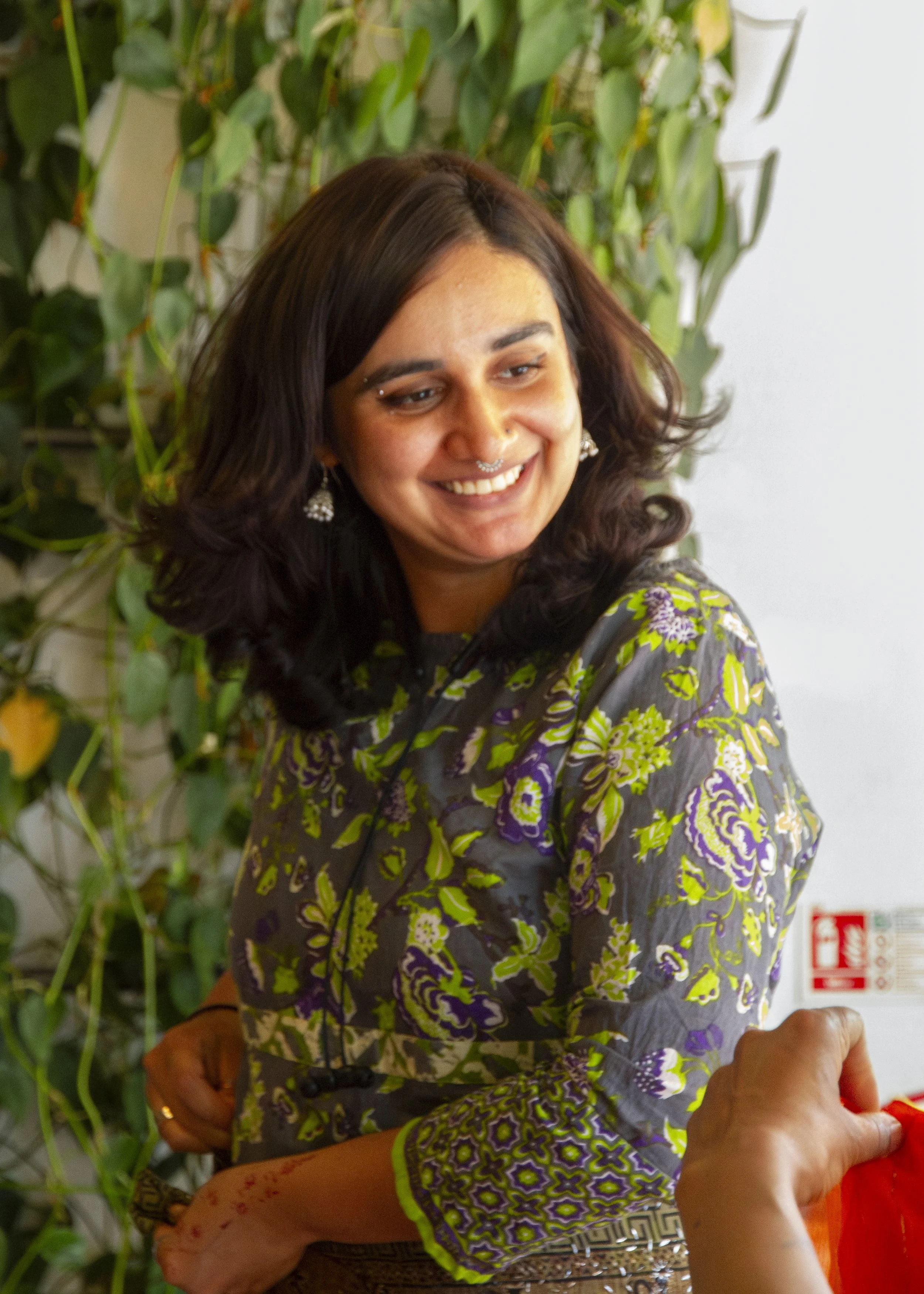 A south Asian woman wearing a green and black top smiles away from the camera, in front of a foliage background