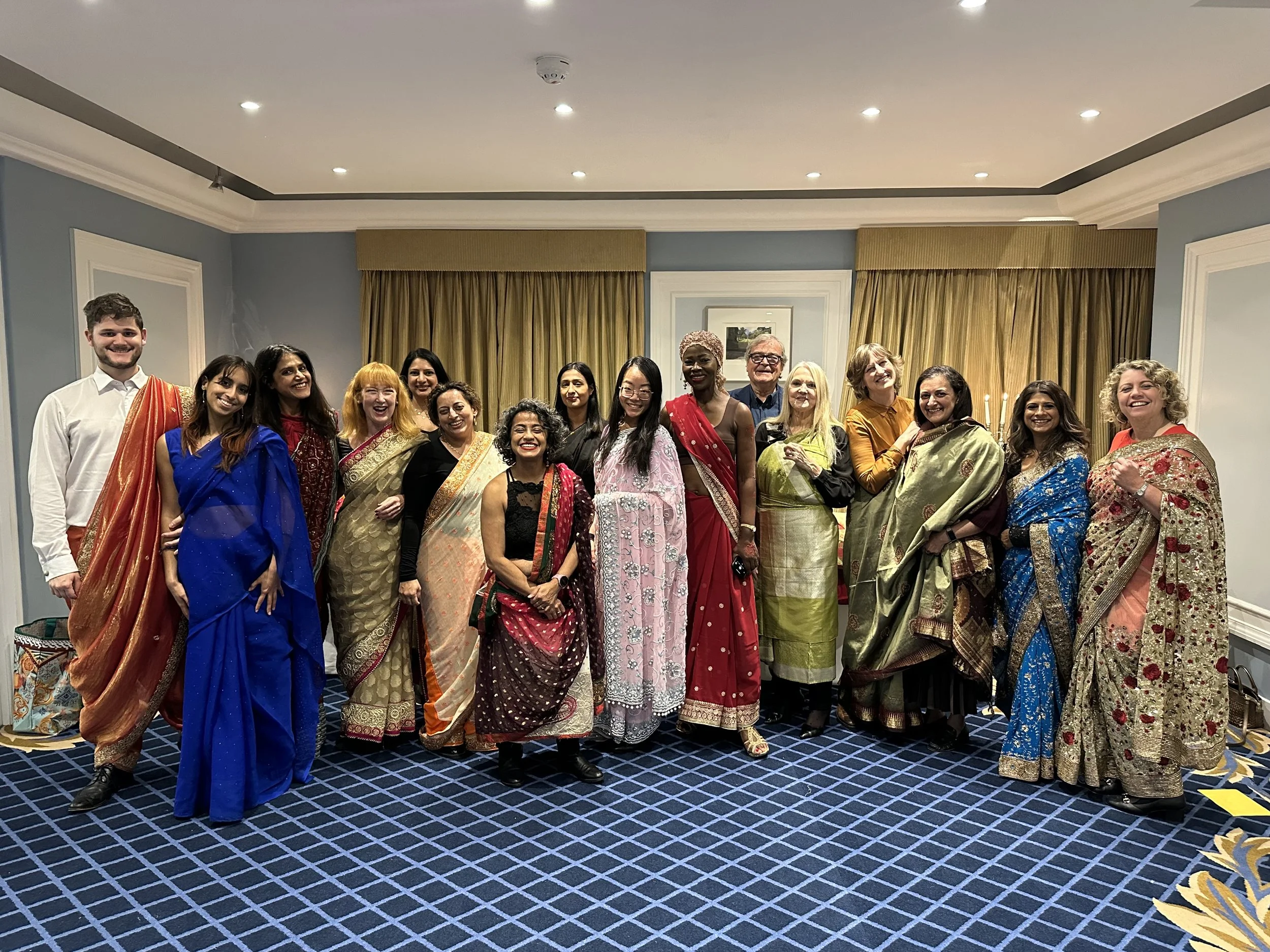 A group of people draped in different coloured sarees crowd together smiling at the camera