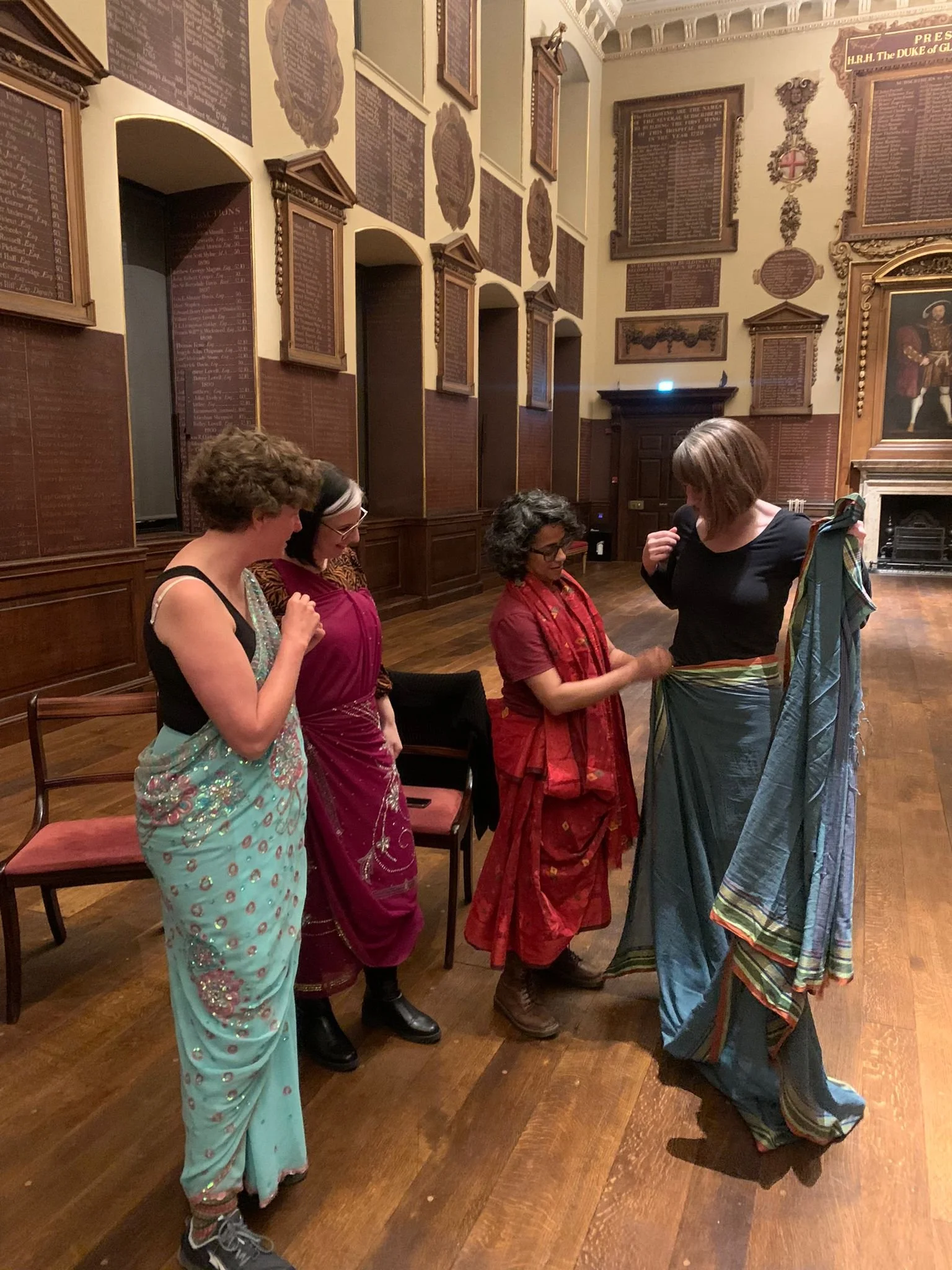 Poppy helps a woman drape herself into a grey saree, while two other women watch