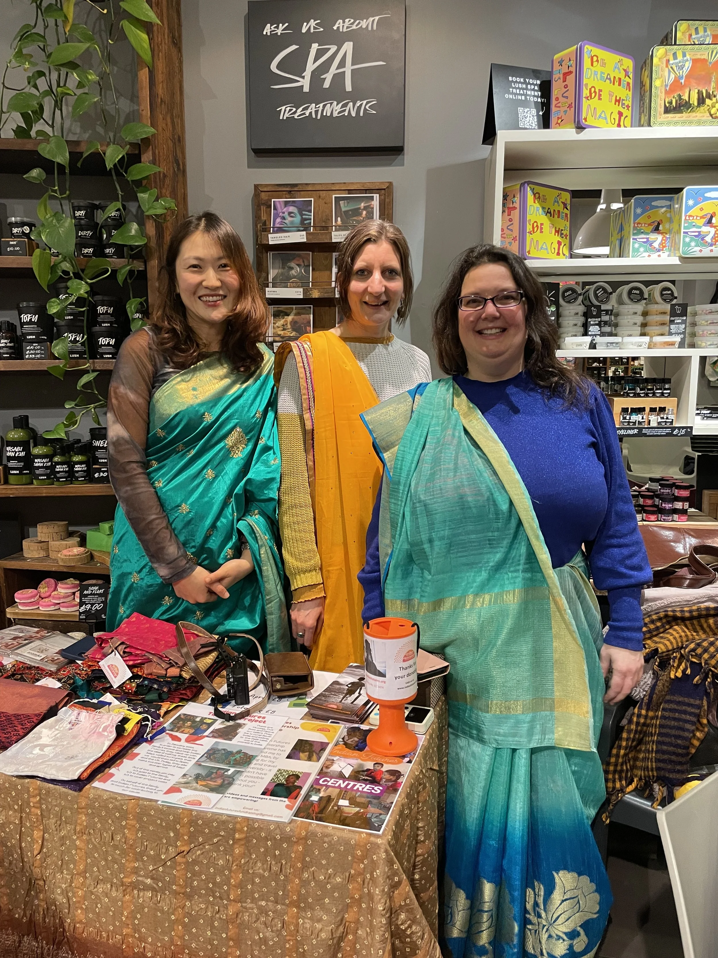 Three women wear colourful sarees behind a table and smile