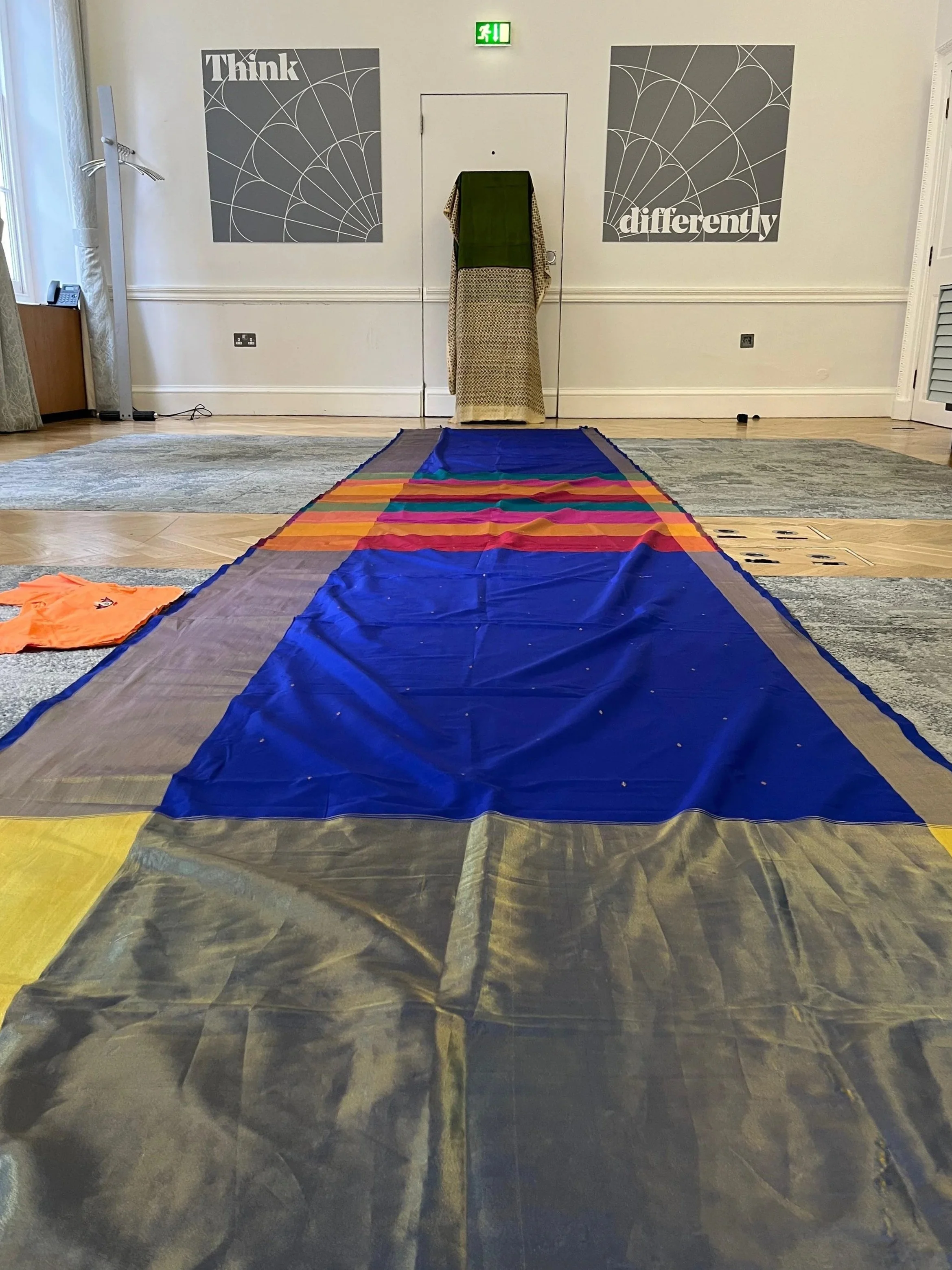 A blue colourful stripy saree is laid on the floor. The wall behind it has two posters, one that says think and the other says differently. Between them is a green patterned saree hanging on a rectangular post
