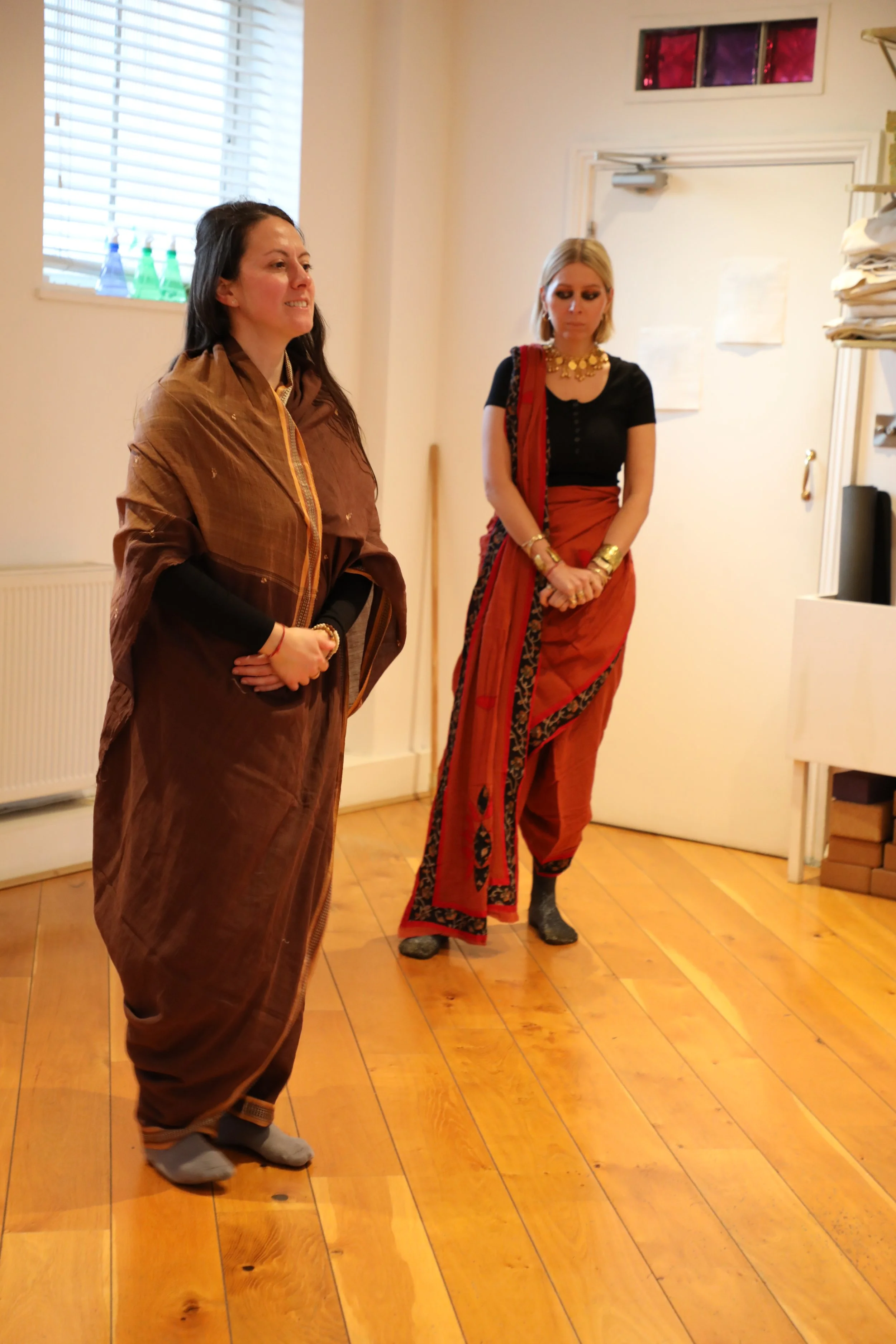 Two women stand together wearing sarees in the trouser drape. Not looking at the camera