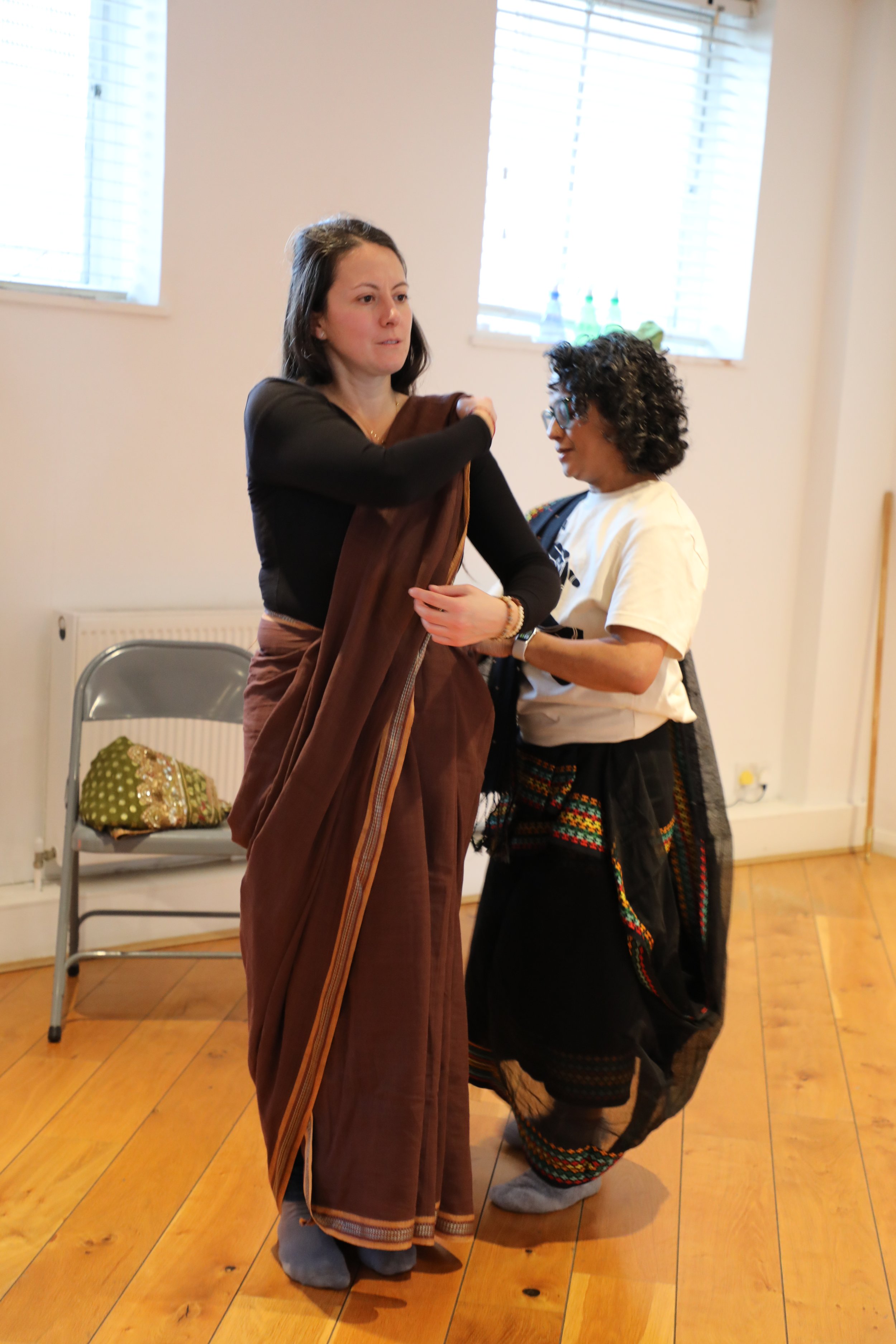 A woman wearing a white t shirt and black saree helps another woman drape a brown saree on herself