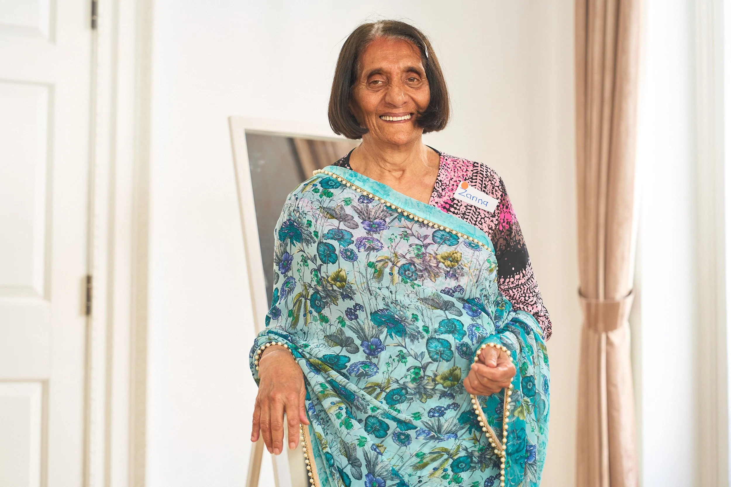 An elderly South Asian woman wearing a patterned blue saree smiles toward the camera