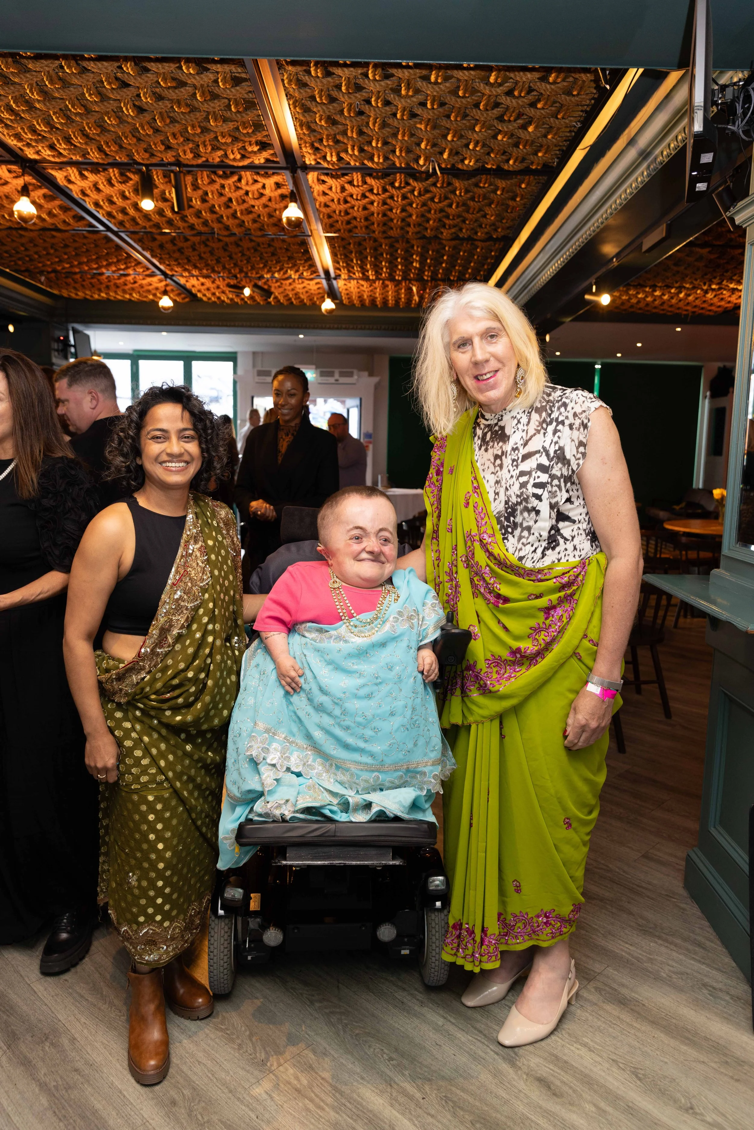 Two people draped in sarees stand next to a third person sitting in an electric wheelchair also draped in a saree