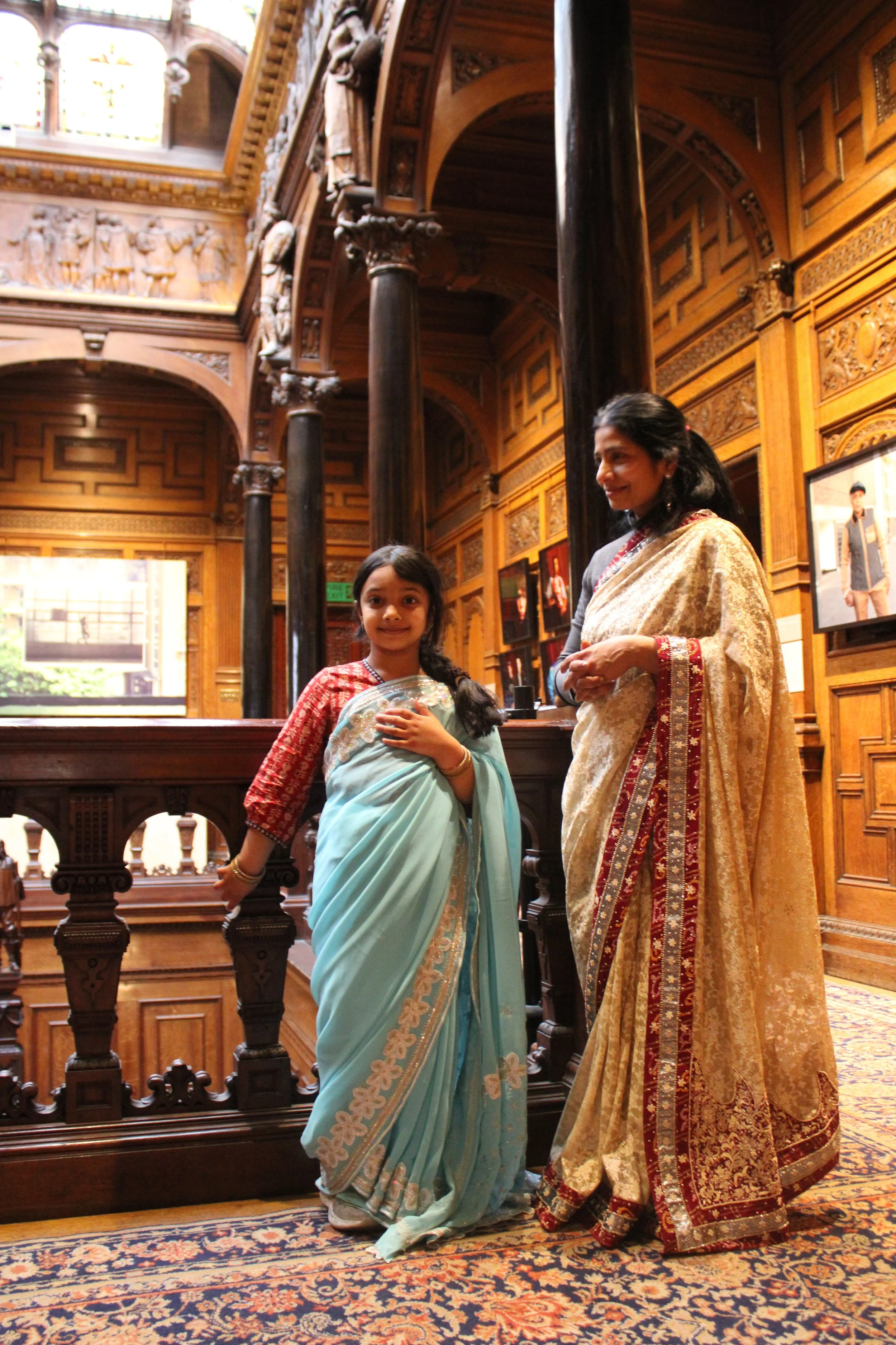 A south asian mother and daughter draped in sarees stand in the foreground in front of a bannister