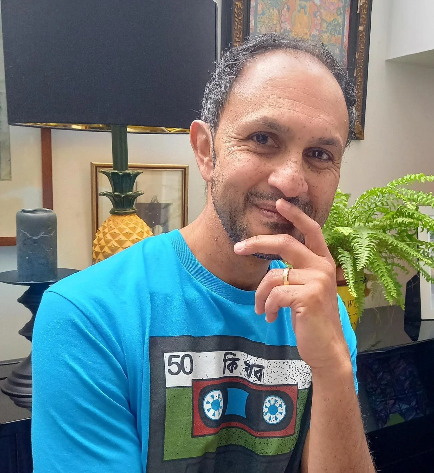 Tauhid as a brown man wearing a blue Kee Khobor logo (cassette player) t-shirt he is smiling closed mouth and his hands are slightly covering his lips.