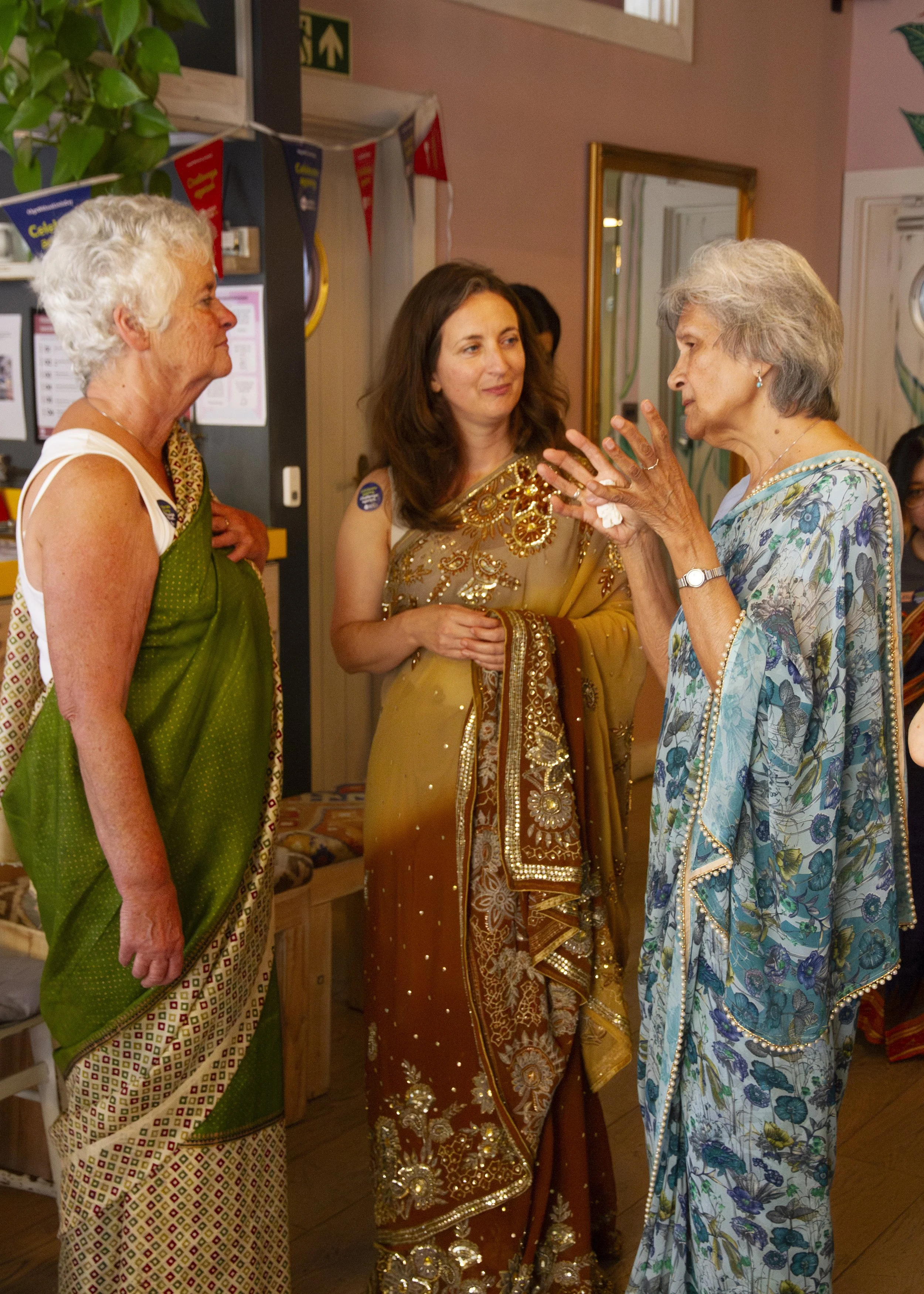Three women wearing sarees stand together and converse