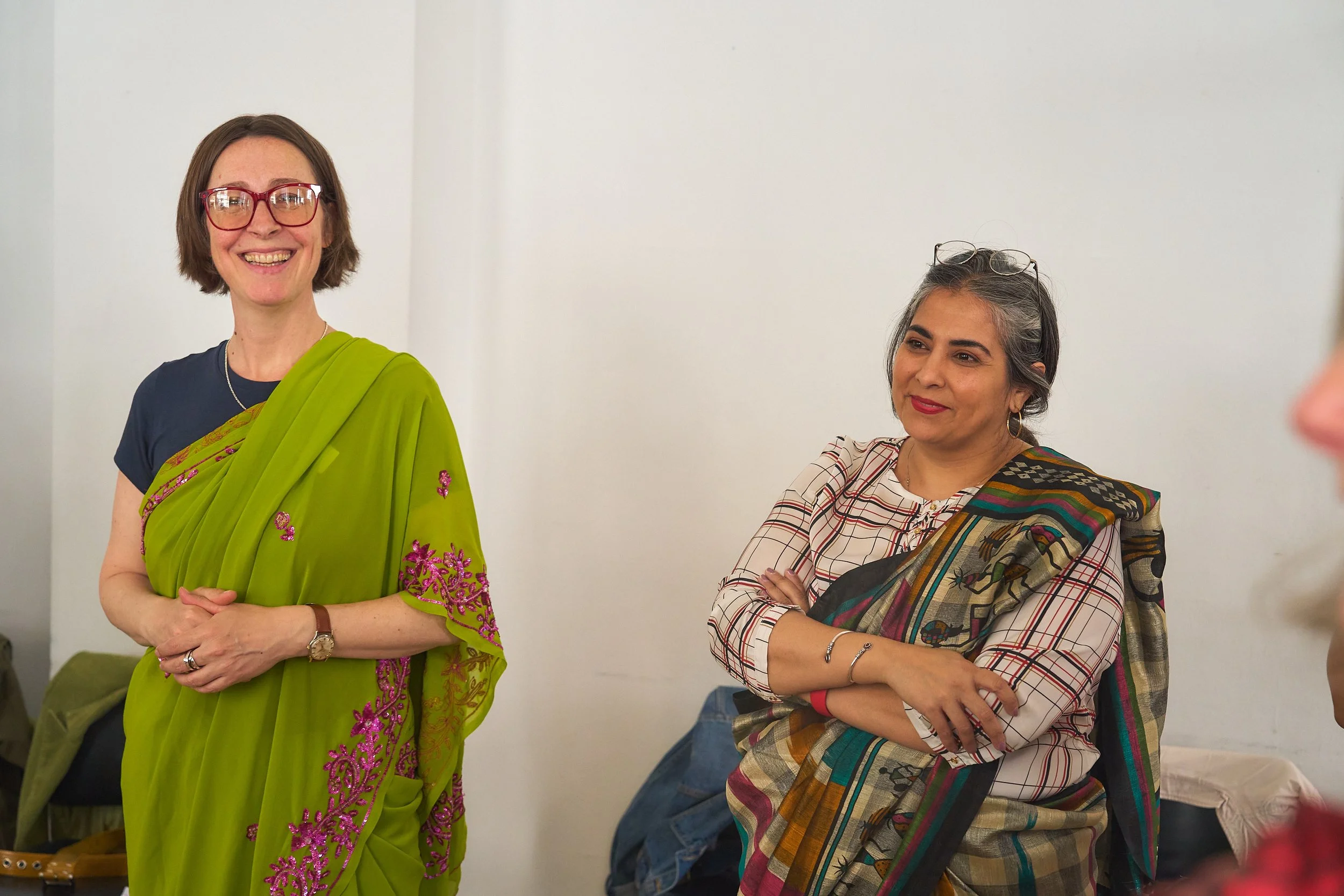 Two women wearing sarees smile, one looking at the camera and one looking at something outside of the shot