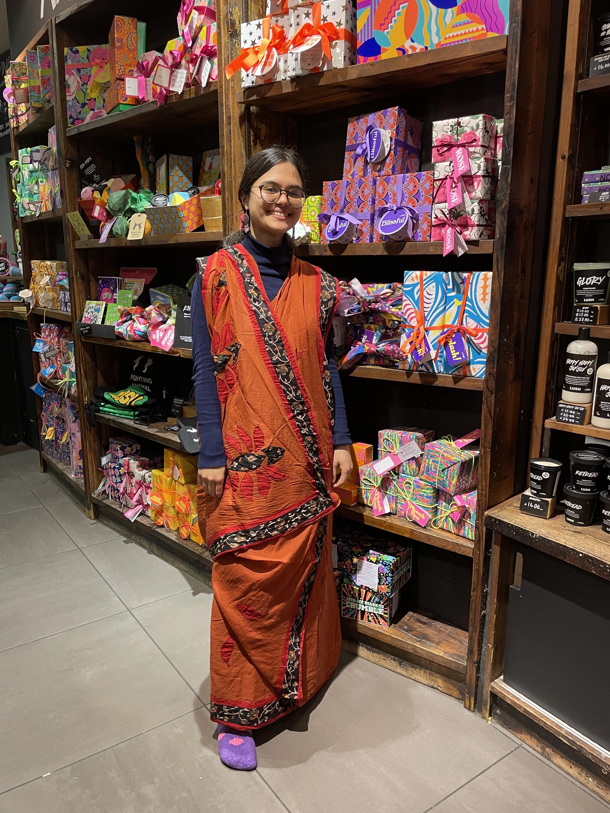 A woman wearing an orange saree smiles and stands in front of a colourful wall