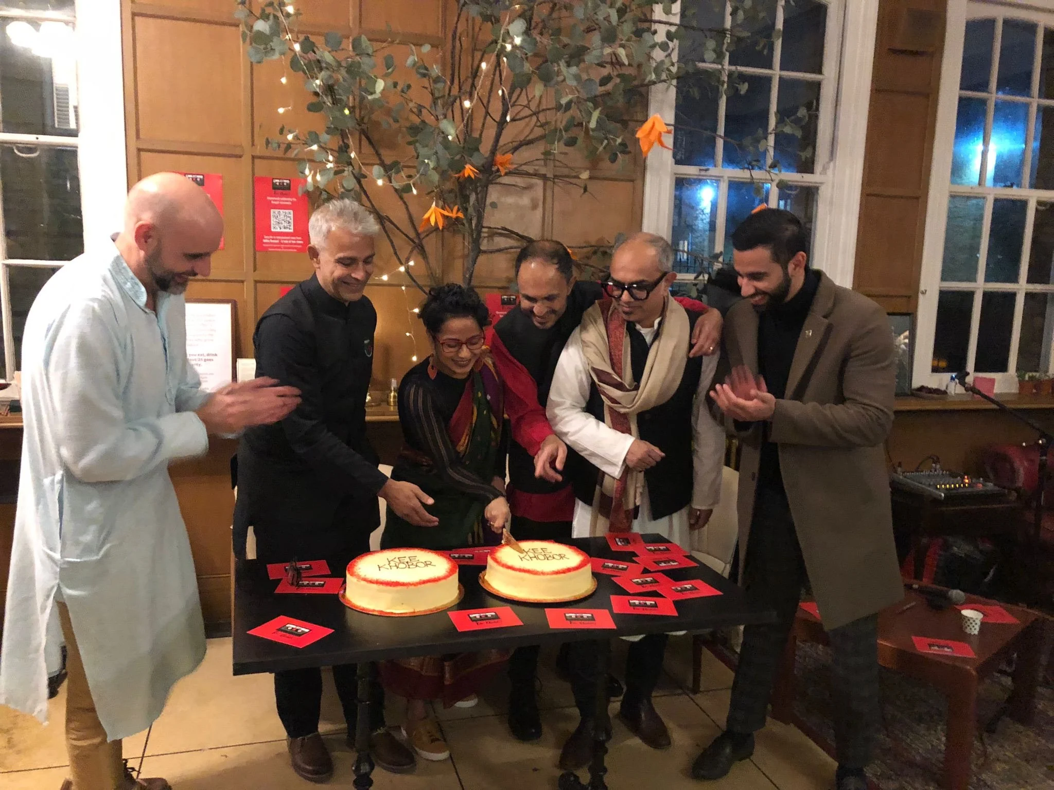 6 people are huddled around the table at the Kee Khobor celebration about to cut a cake they are smiling and have there arms around each other. Poppy the Founder of Re-Balance is holding the knife to cut the cake