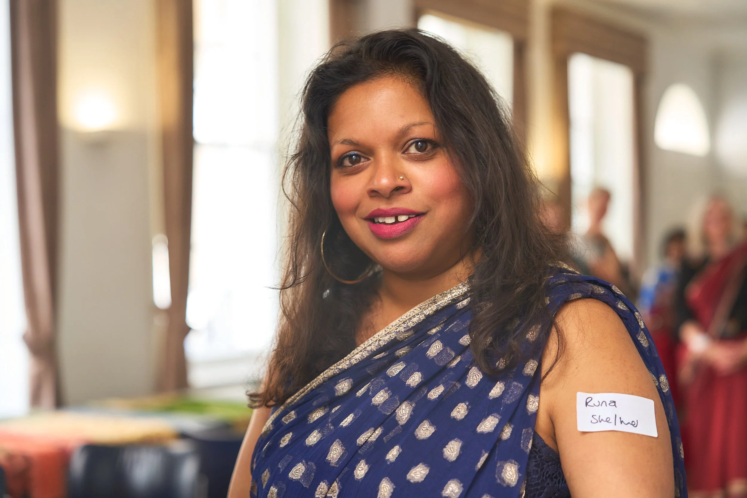 A South Asian woman wearing a deep blue patterned saree smiles at the camera. This is a shot of her upper body and face
