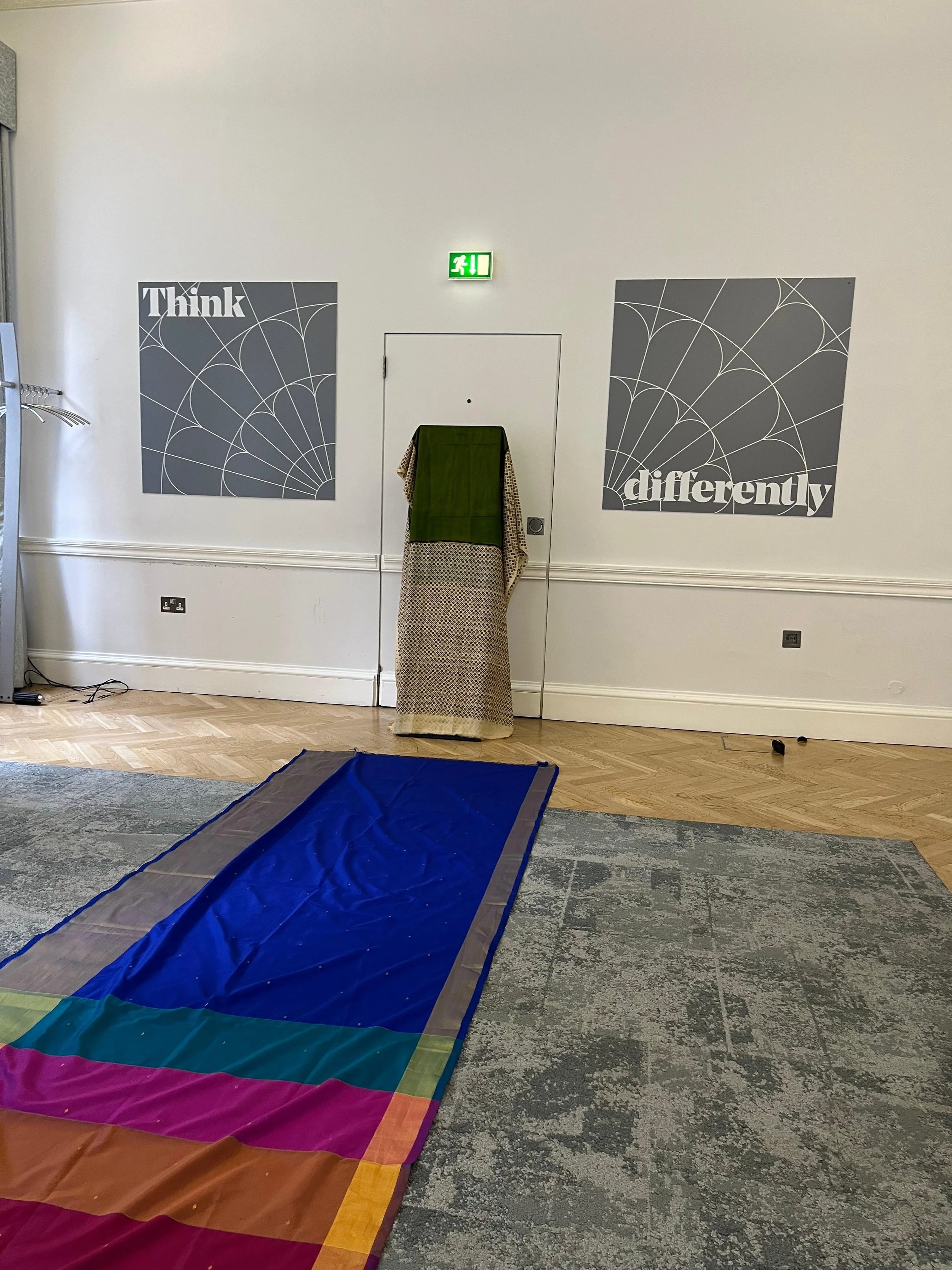 A blue saree laid out on the floor with a green patterned saree hung on a rectangle post behind it