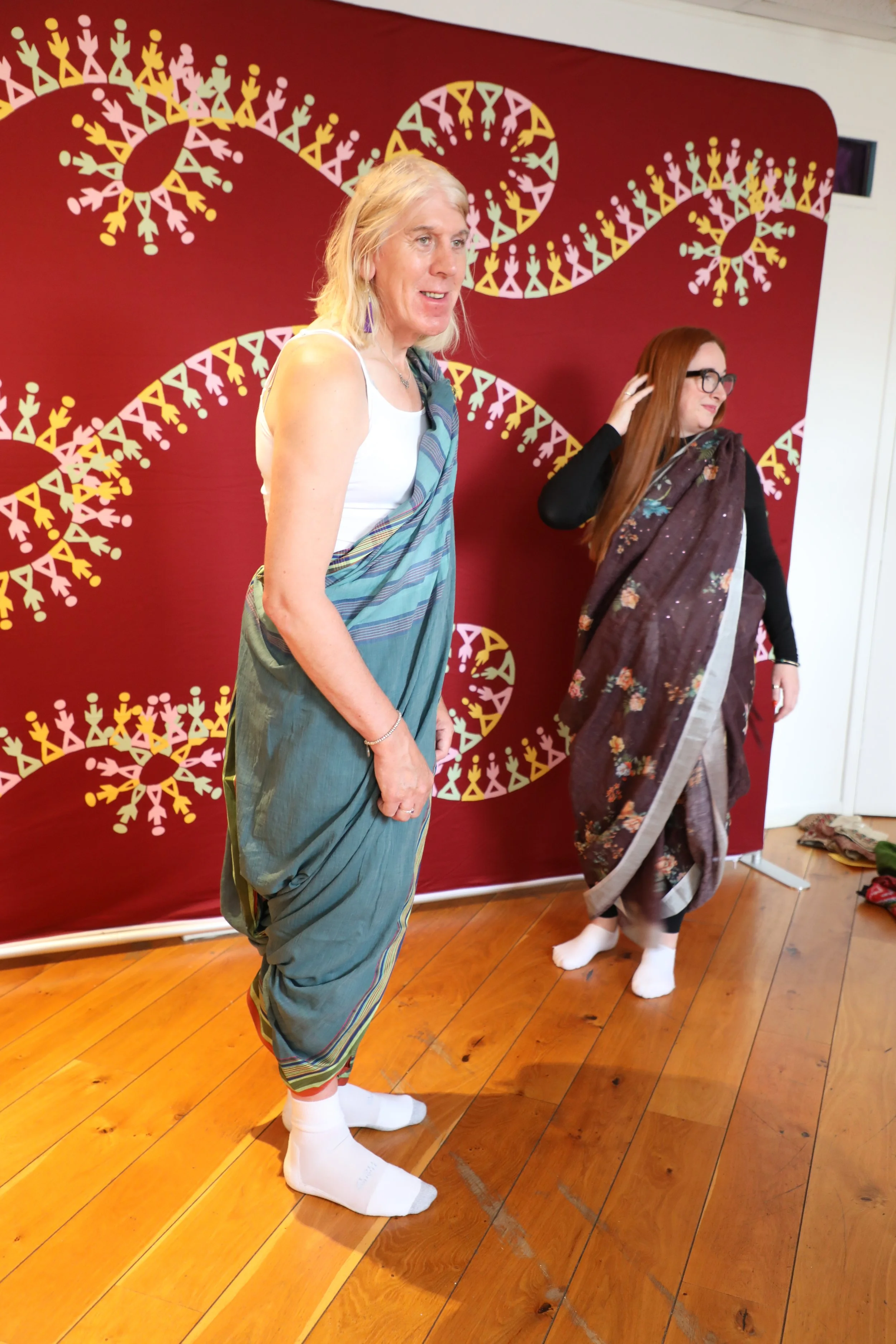 Two people wearing sarees in a trouser drape stand in front of a colourful patterned backdrop - candid shot, they are not looking at the camera