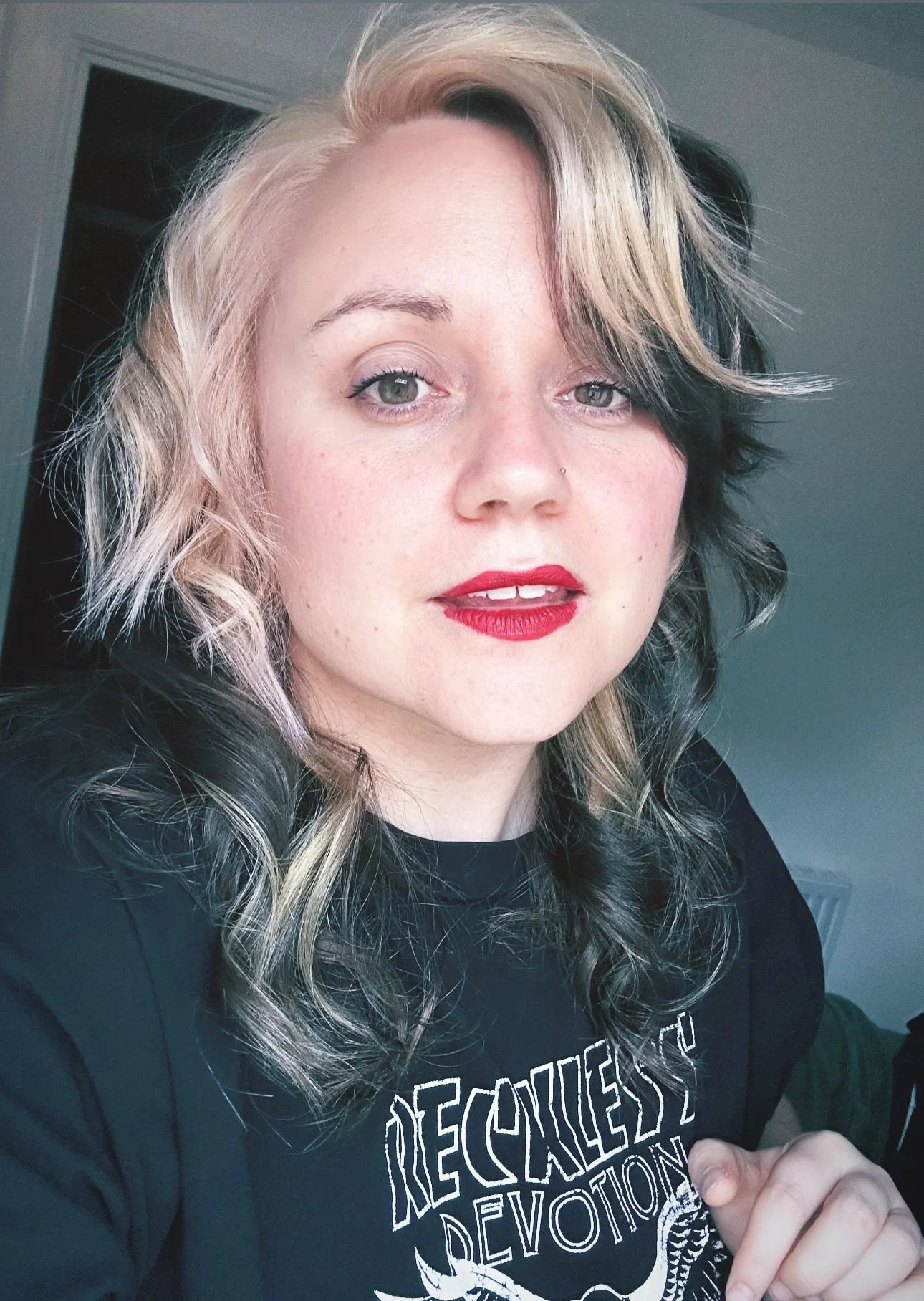 Emma a white non-binary person with blonde and black curly styled hair is wearing red lipstick and smiling open mouthed at the camera.
